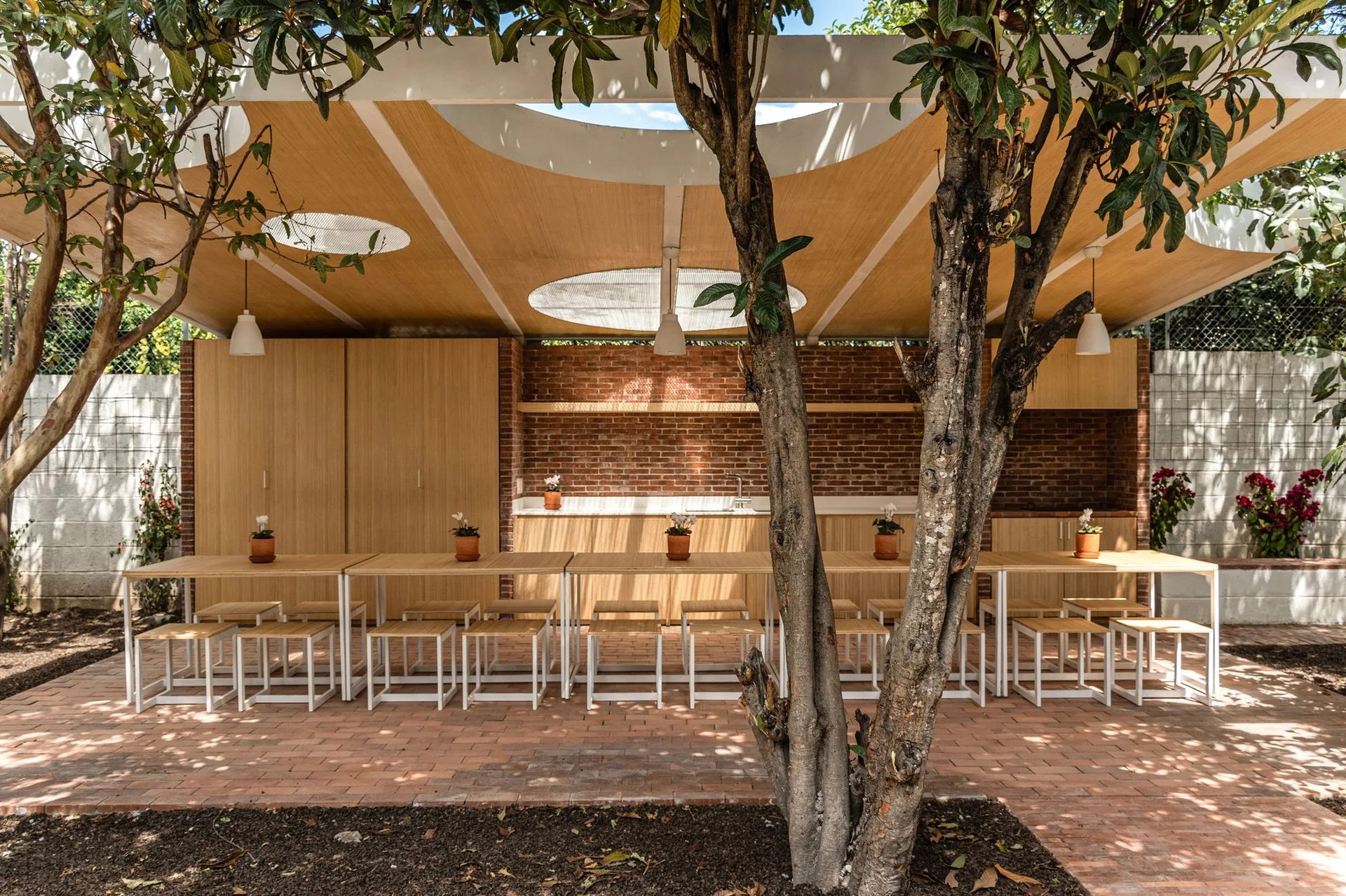 A covered outdoor patio dining area features light wood tables, matching stools, exposed brick walls, and a wooden canopy ceiling with circular cutouts.