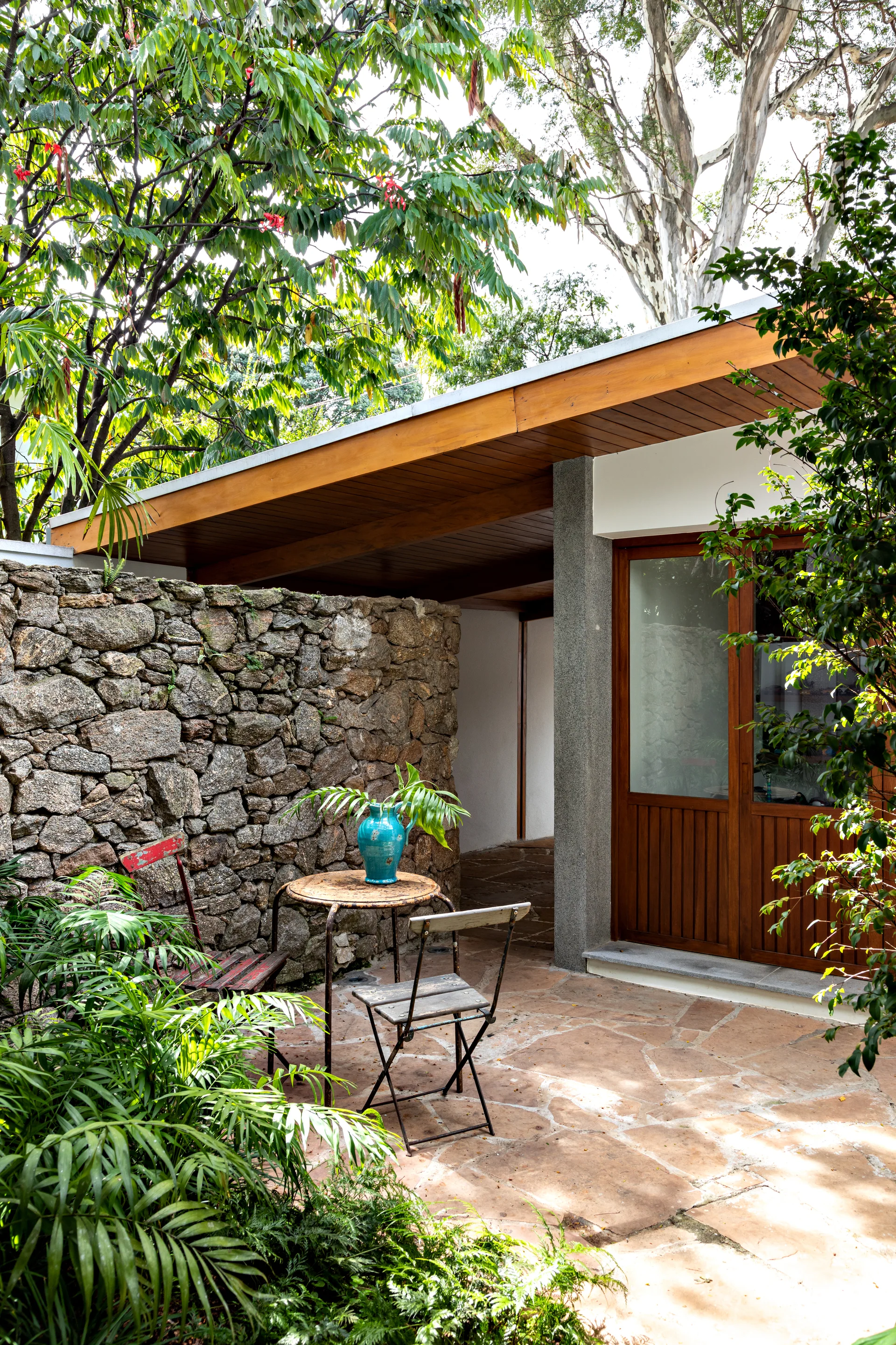 Rustic patio with a stacked stone wall, flagstone flooring, a wooden ceiling overhang, and a small bistro table with two folding chairs.