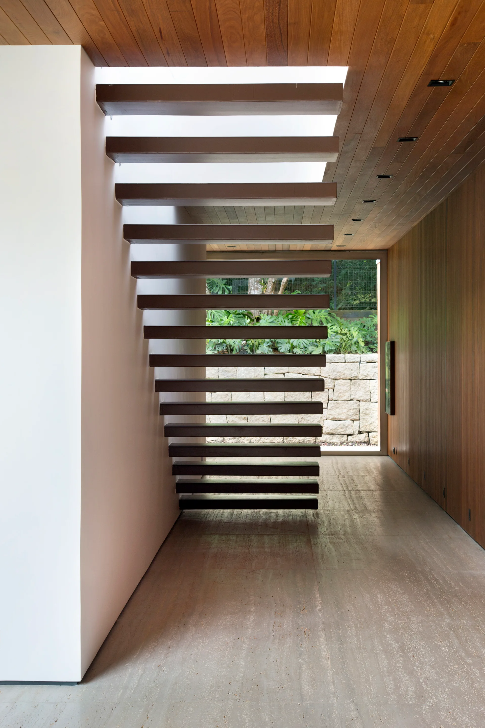 A modern hallway with a cantilevered wooden staircase, wood-paneled walls and ceiling, stone tile flooring, and a window facing a green garden.