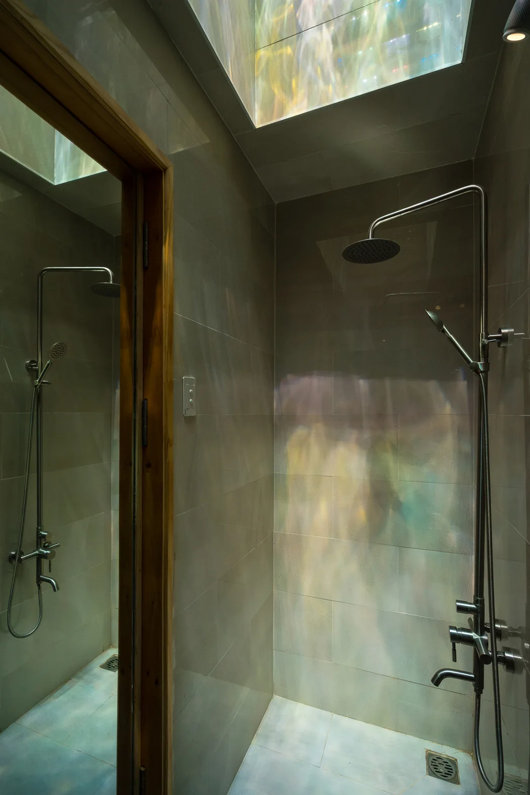 A minimalist shower stall with grey stone tiles and chrome fixtures is illuminated by refracted colorful light from a skylight above.