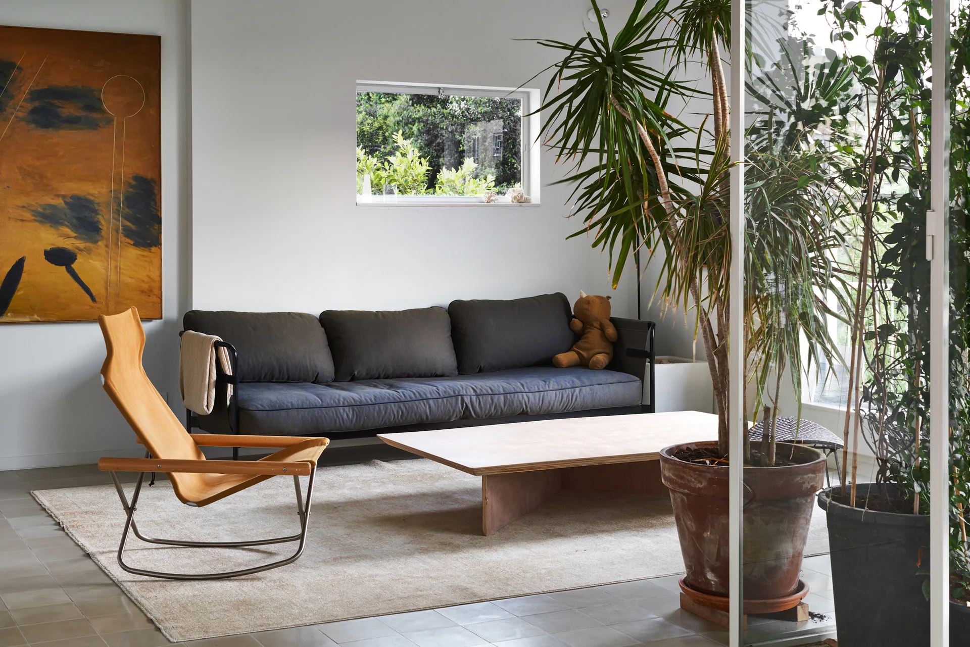 A minimalist living room features a dark grey sofa, a low wooden coffee table, a tan rocking chair, and a large potted plant.
