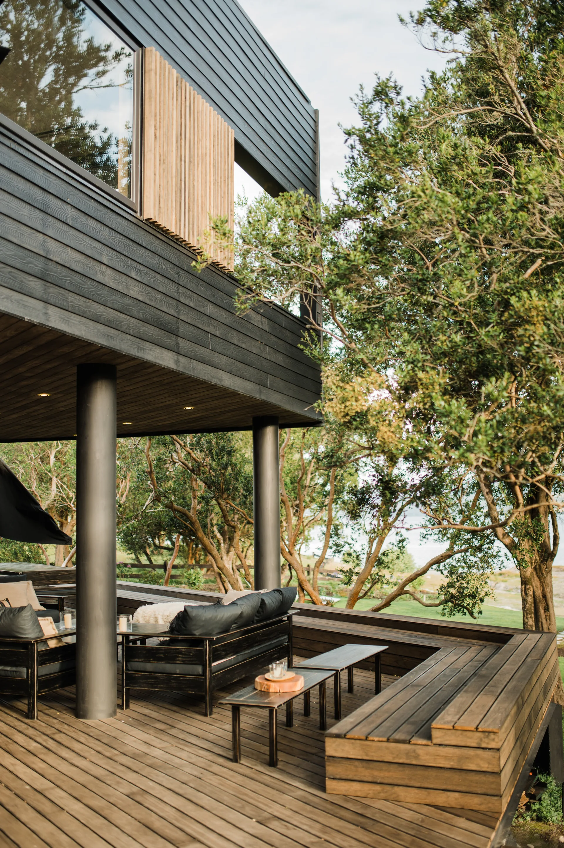 A dark stained wooden deck under a house overhang features black outdoor sofas, a built-in wooden bench, and structural metal columns near green trees.