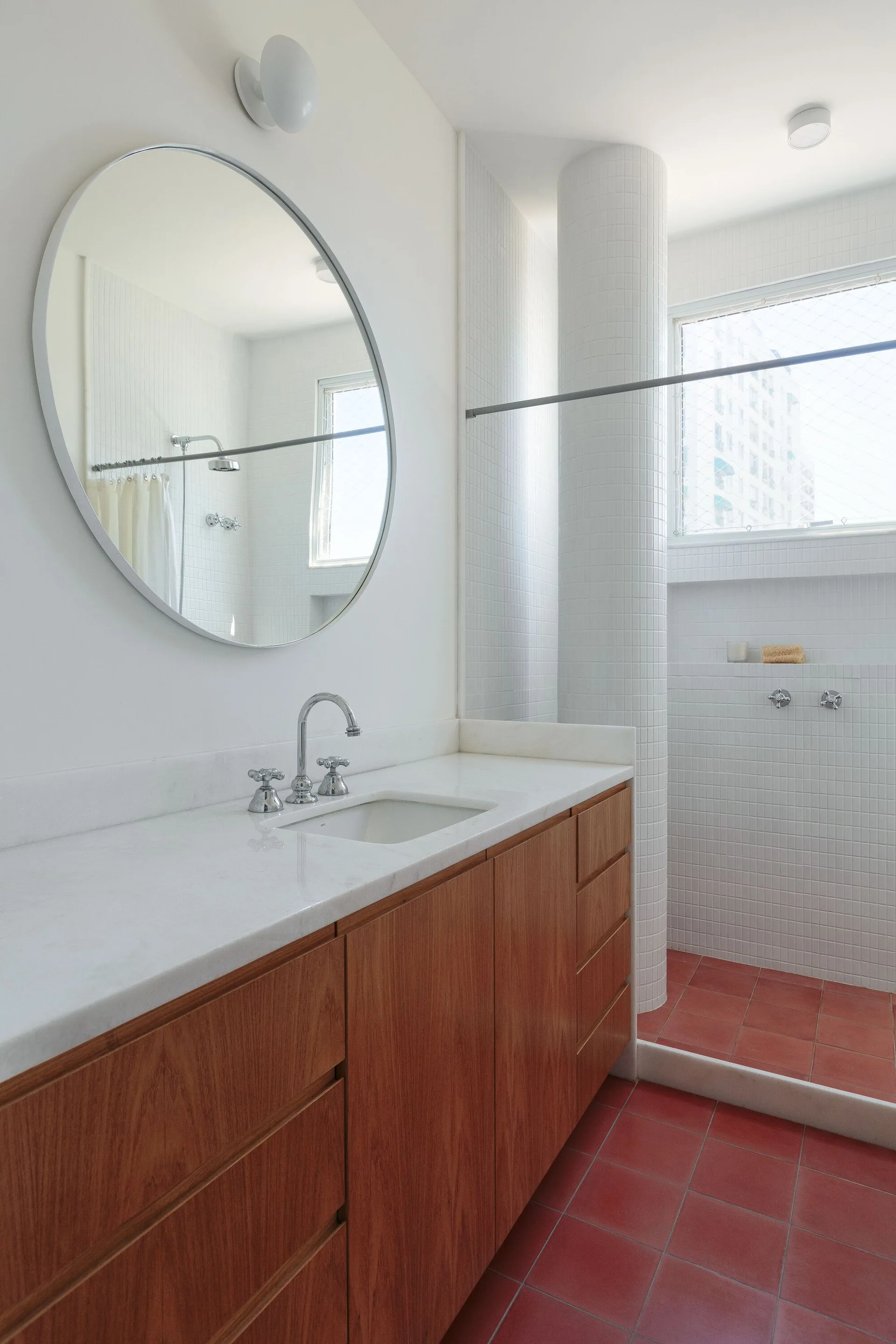 A modern bathroom features a rich wood vanity with a white countertop under a round mirror next to a shower with white mosaic tile and red tiled floor.