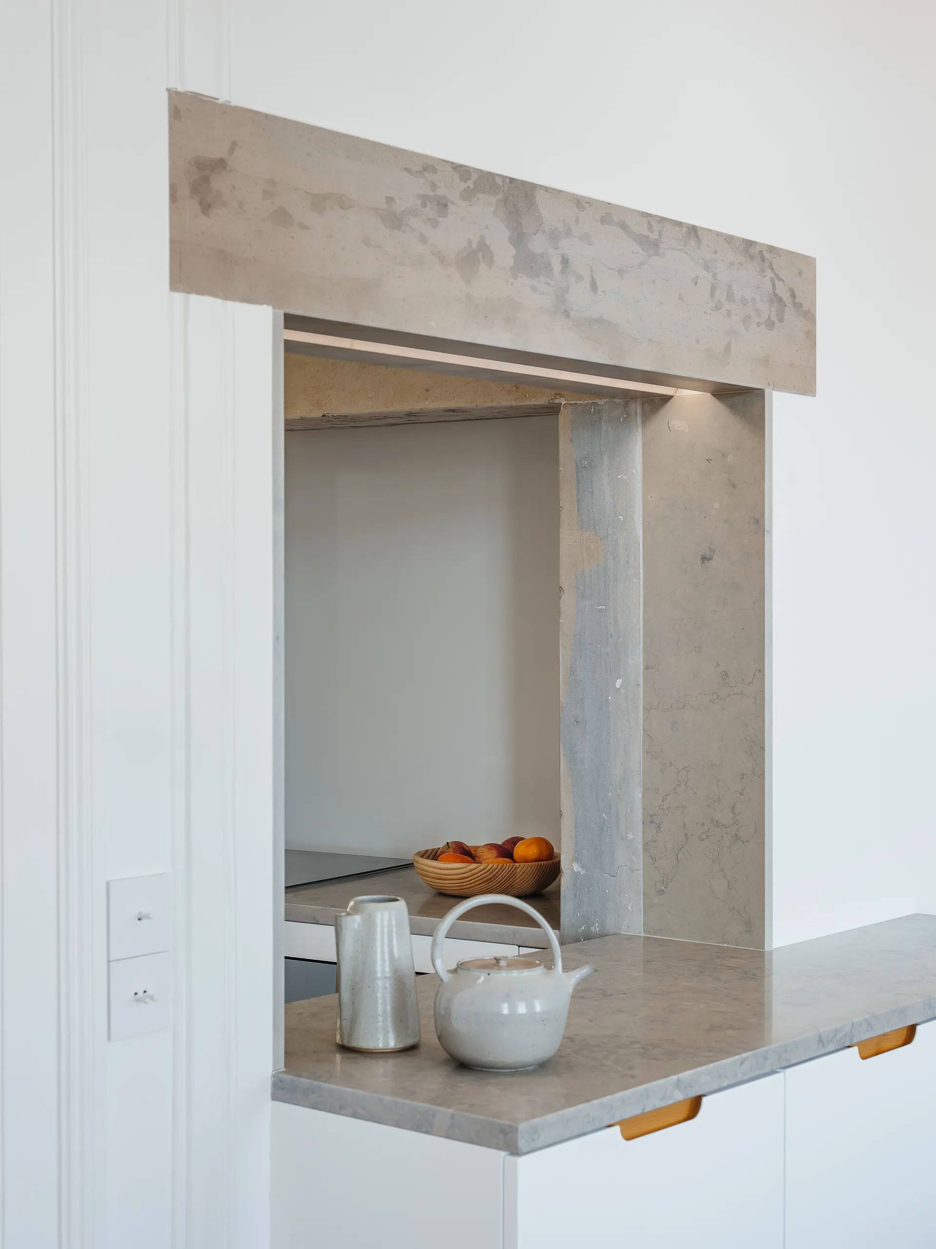 A minimalist kitchen pass-through framed in grey stone features a matching countertop, white cabinets, and ceramic tableware.