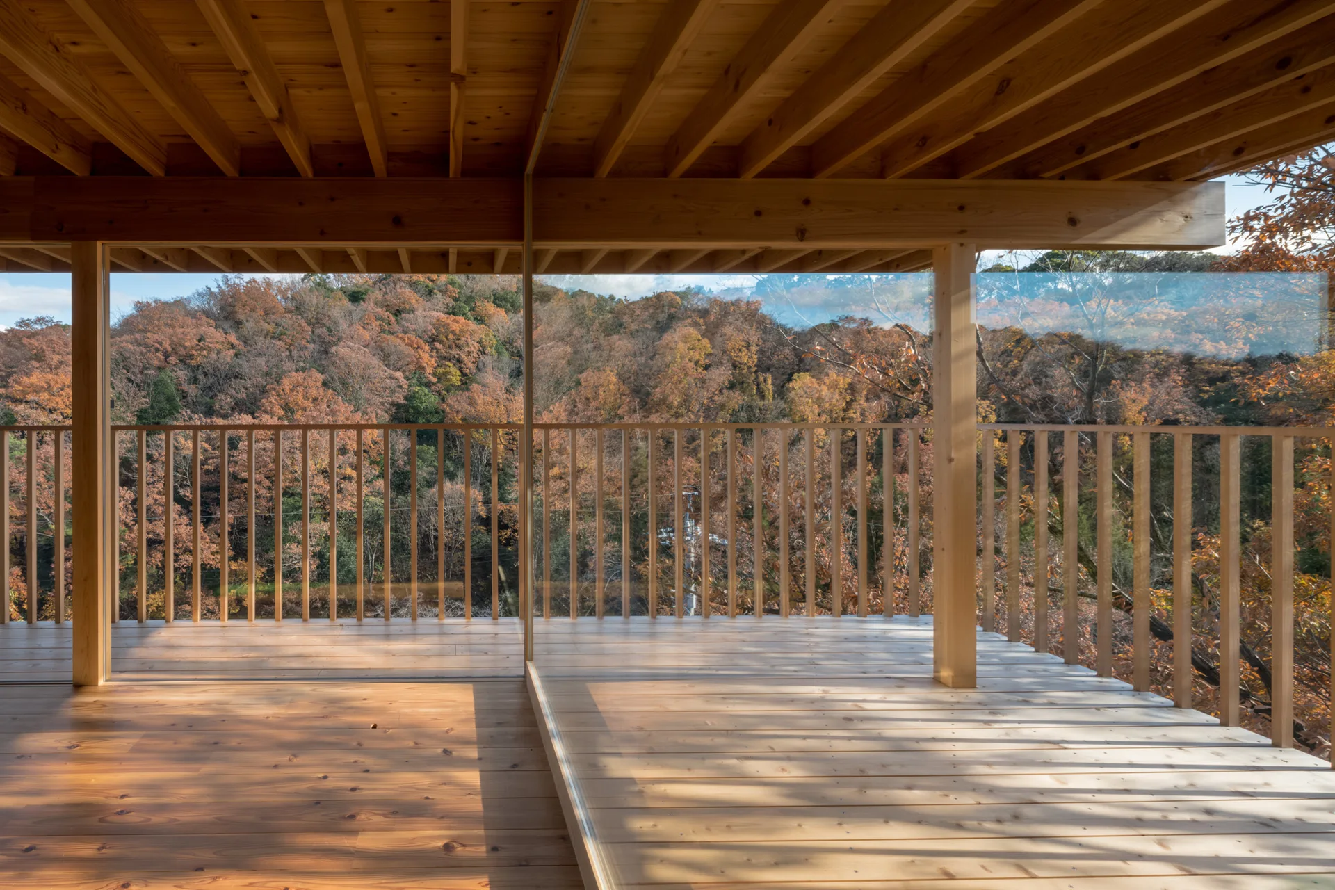 A wooden balcony with an exposed timber ceiling and vertical slat railing overlooks a dense forest with autumn foliage.