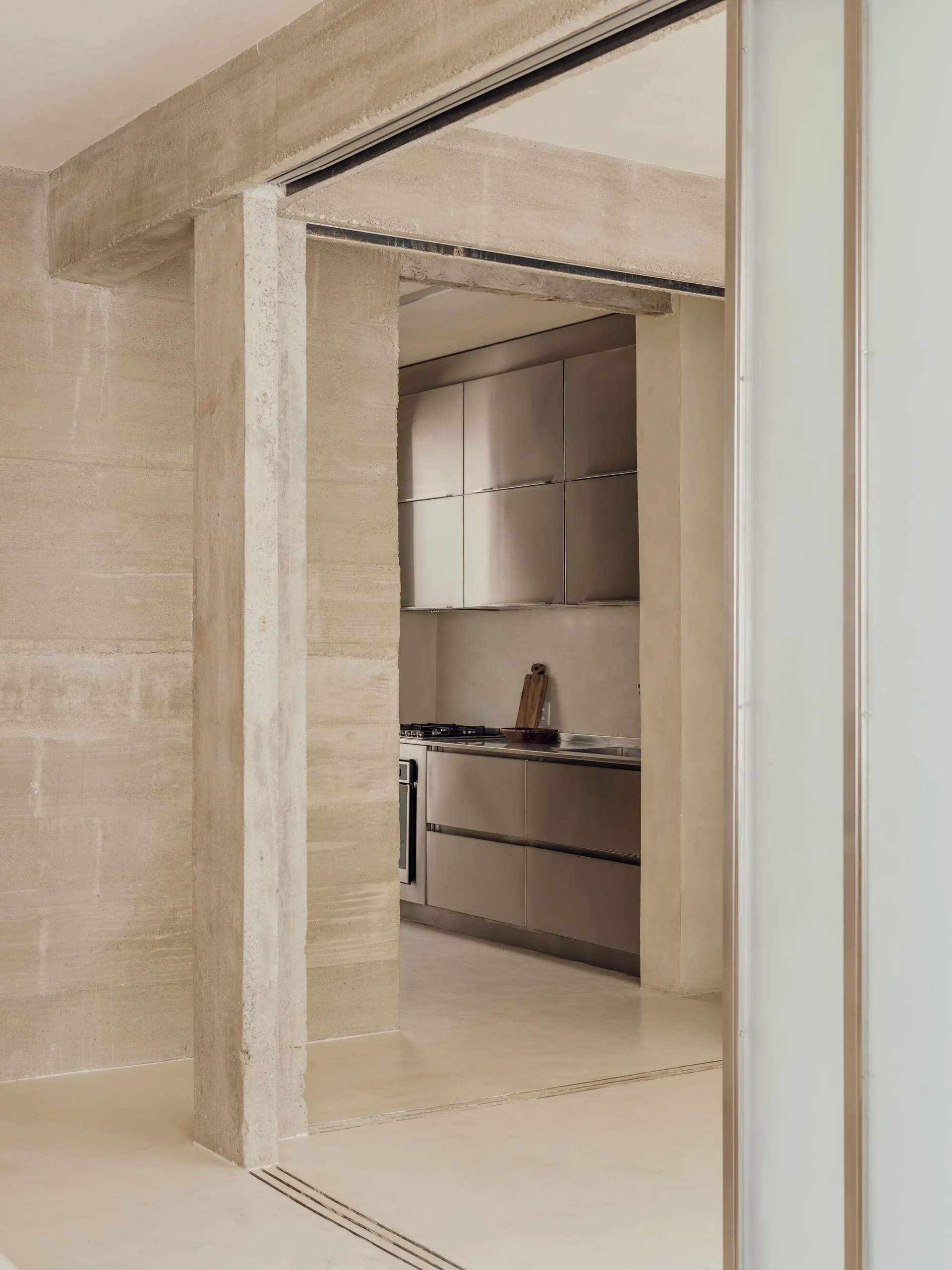 A minimalist kitchen featuring brushed stainless steel cabinetry is framed by textured raw concrete walls and structural pillars.