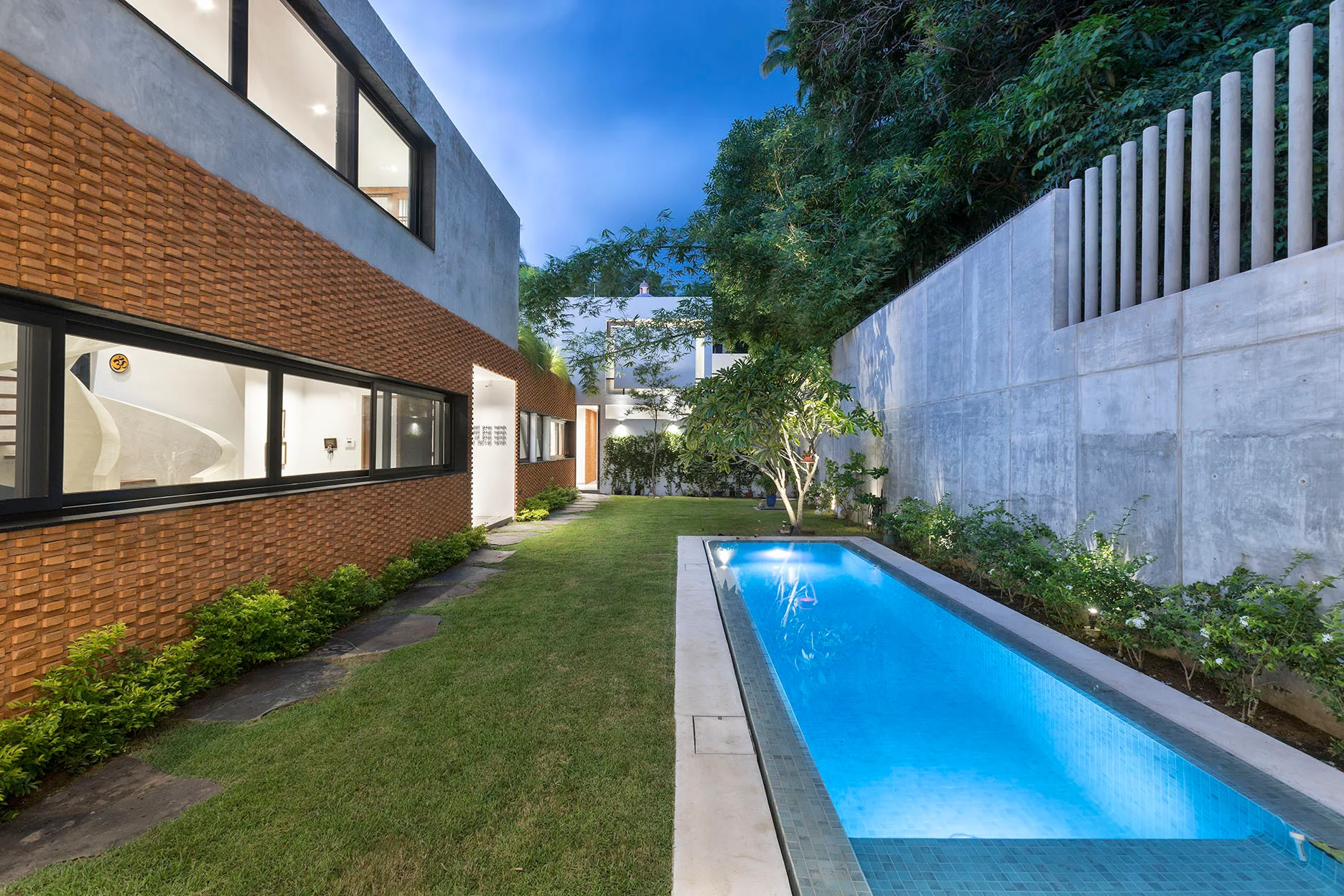 A long rectangular swimming pool glows blue at dusk in a grassy yard between a textured brick house and a tall concrete boundary wall.