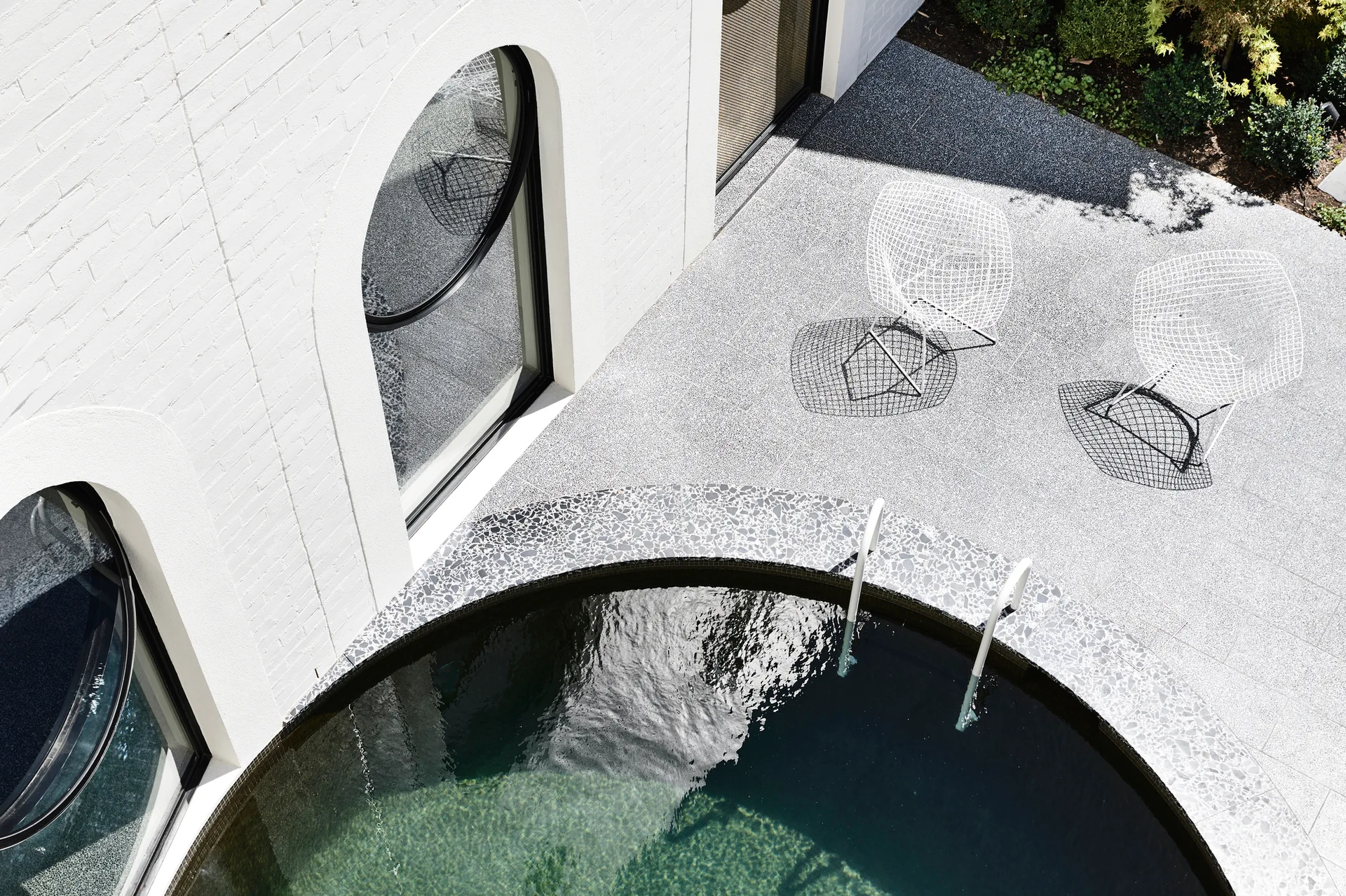 An overhead view of a circular swimming pool next to a white brick wall with arched windows and two white wireframe chairs on a grey patio.