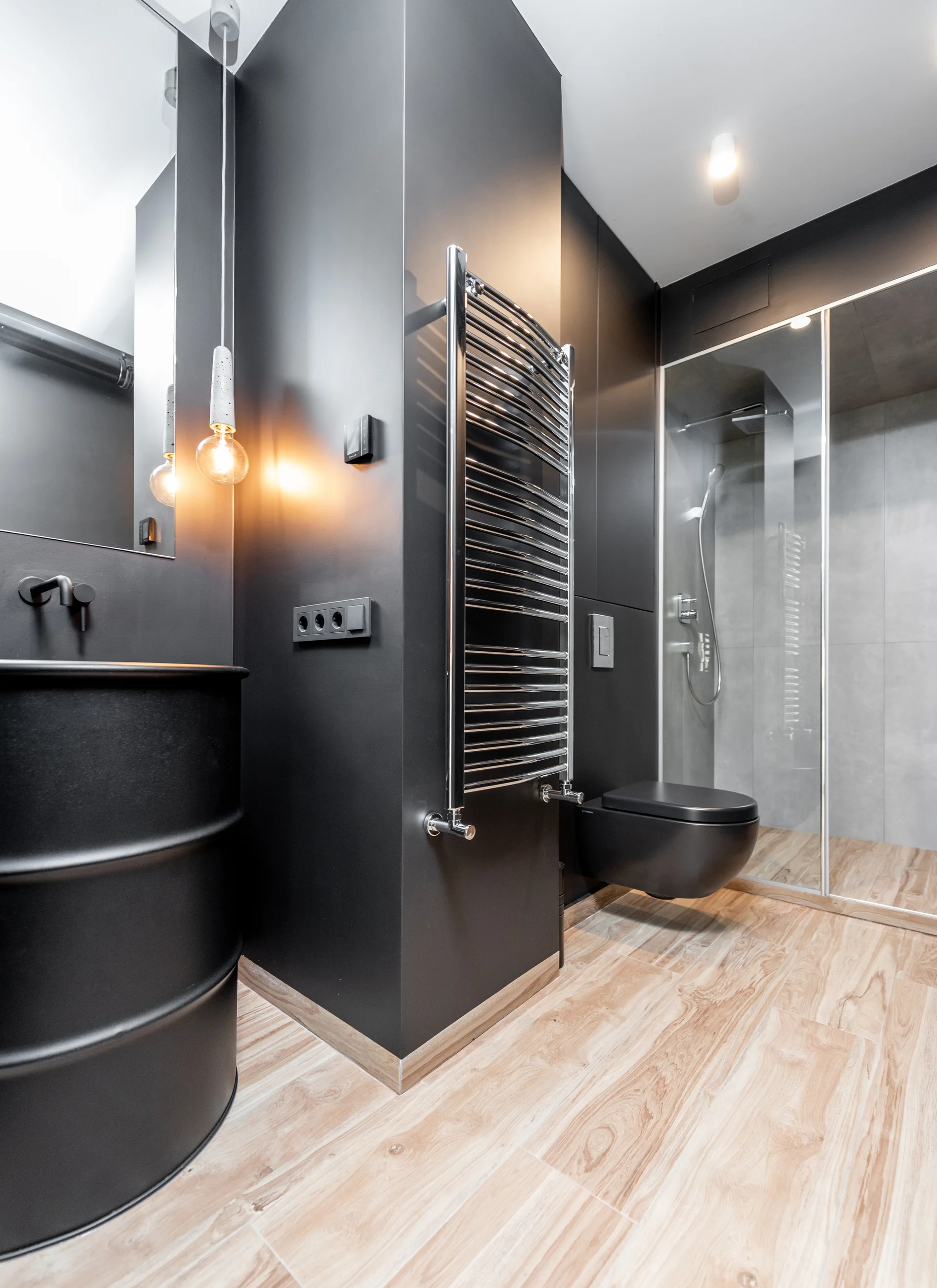 A modern bathroom features matte black walls, a wall-hung black toilet, a chrome towel warmer, and wood-look floor tiles leading to a shower.