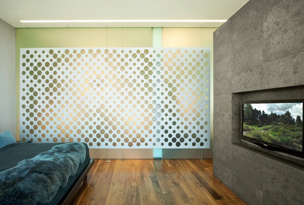 A modern bedroom features a white hanging partition screen with circle cutouts, hardwood floors, and a television recessed inside a grey stone wall.