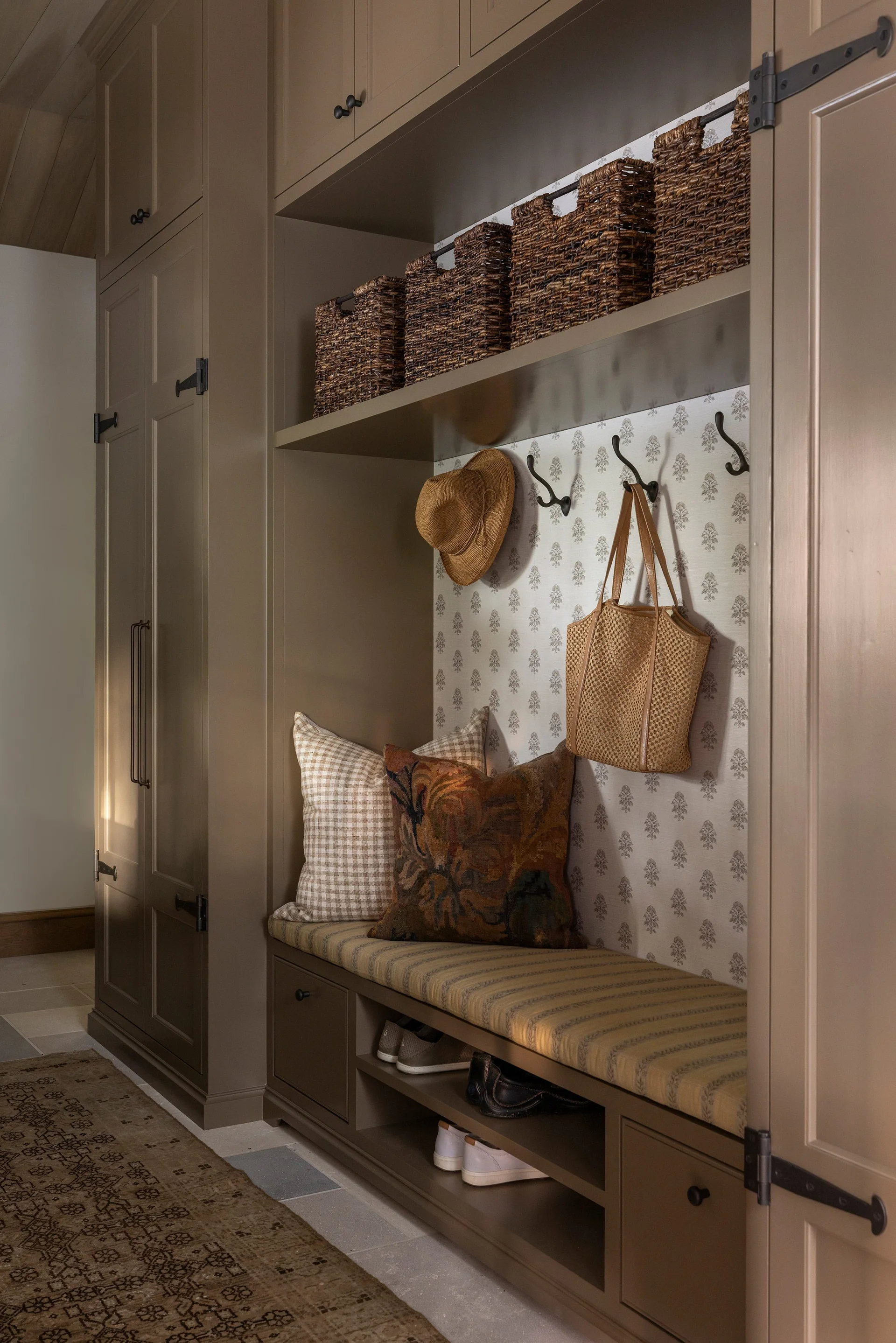 A traditional mudroom with custom taupe cabinetry, built-in bench seating with pillows, floral wallpaper backing, and wicker baskets.