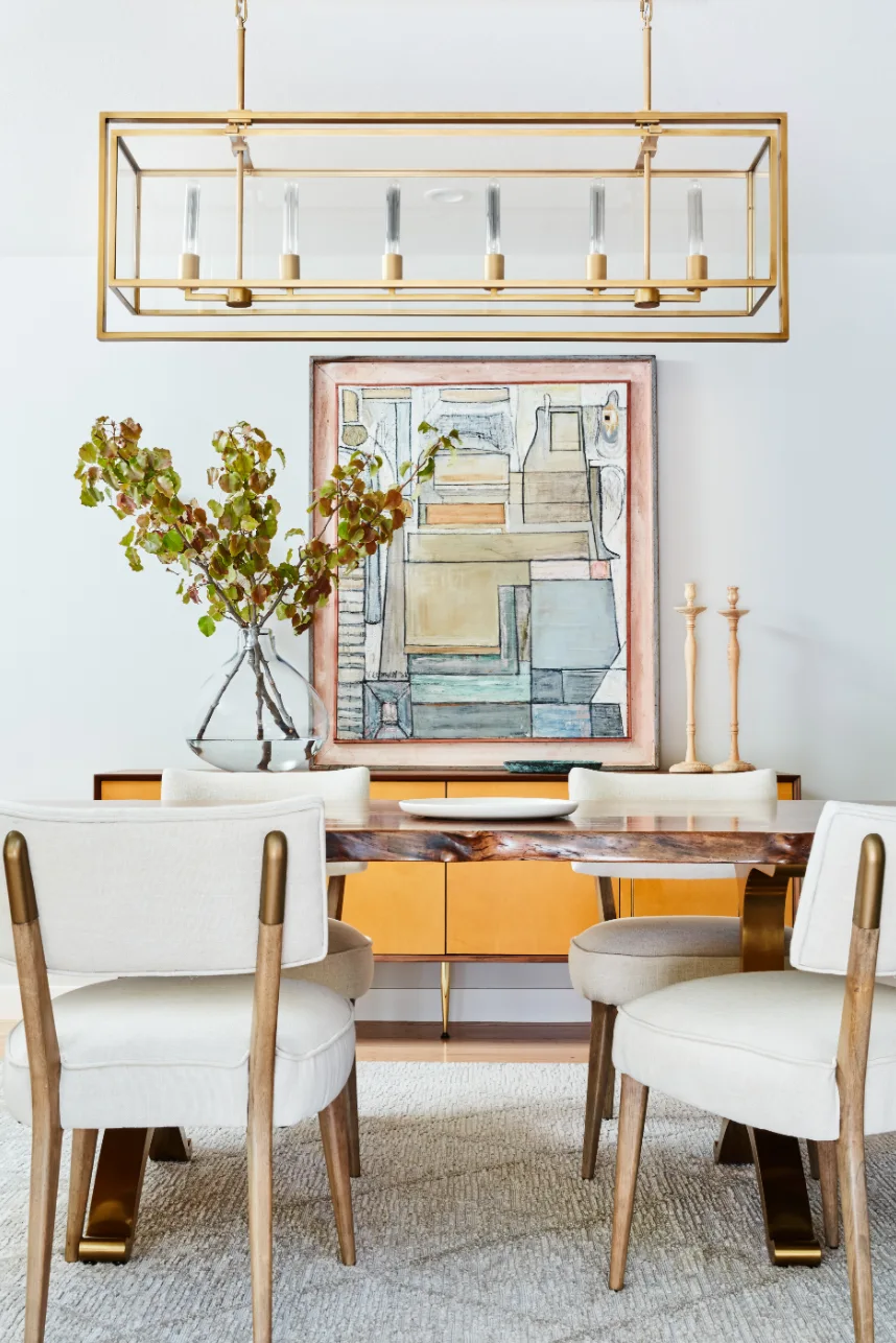 A modern dining room features a live-edge wood dining table, cream upholstered chairs, a vibrant orange sideboard, and a rectangular brass chandelier.