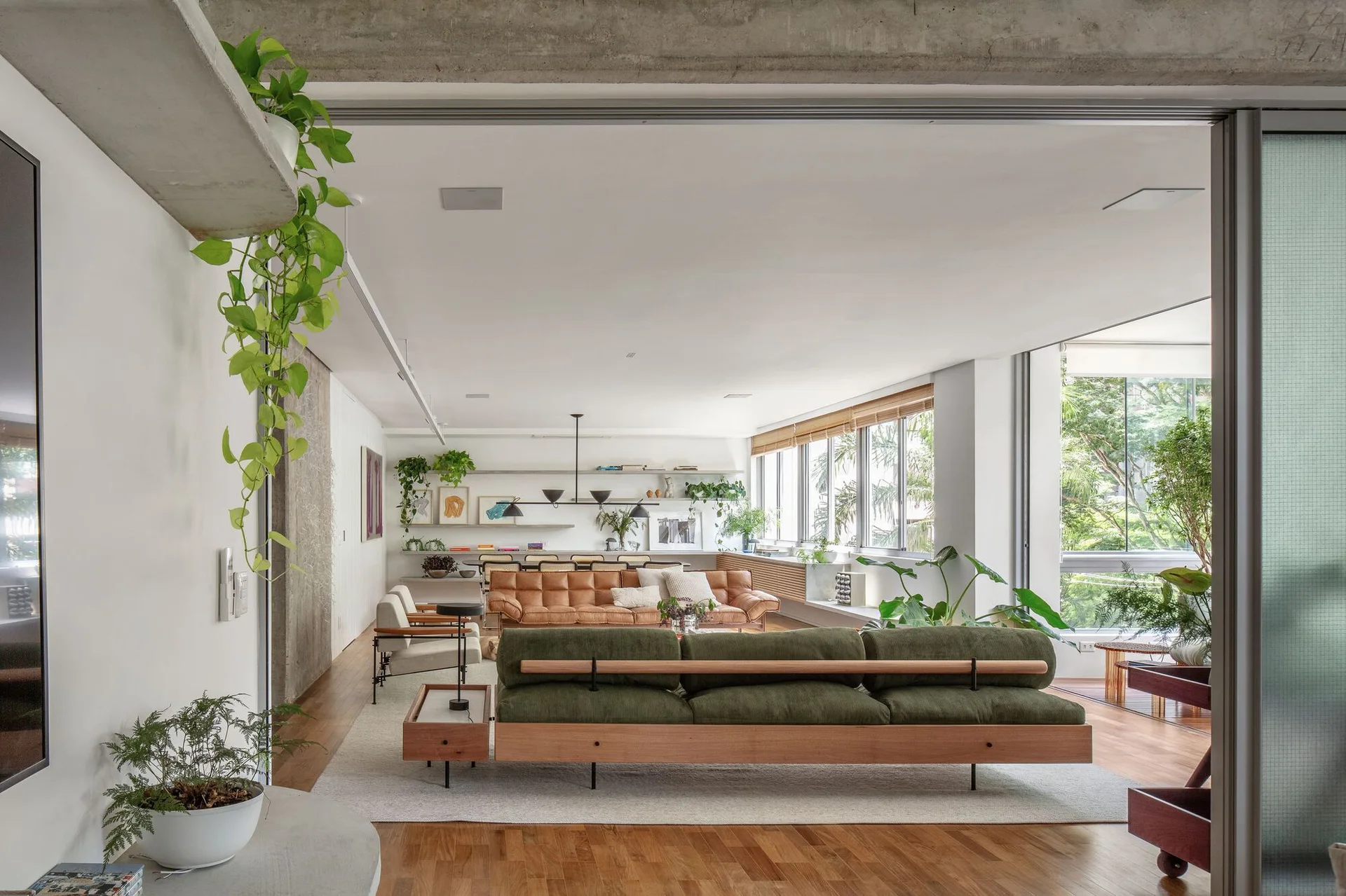 An open living room features an olive green corduroy sofa, a tan leather sofa, wooden floors, and an exposed concrete beam near large windows.