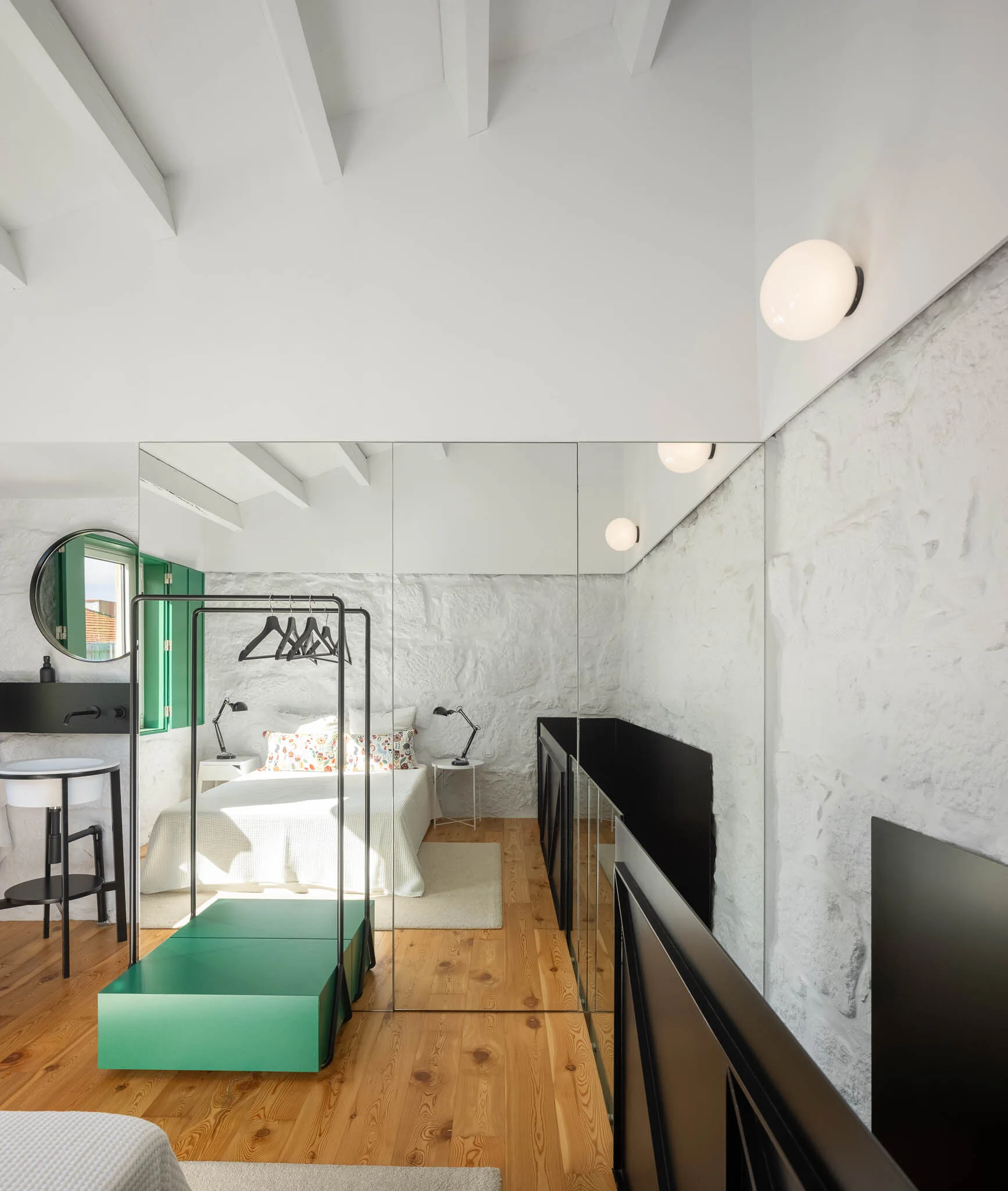 A contemporary bedroom featuring textured white stone walls, exposed ceiling beams, a black clothing rack, and a full-height mirrored partition.