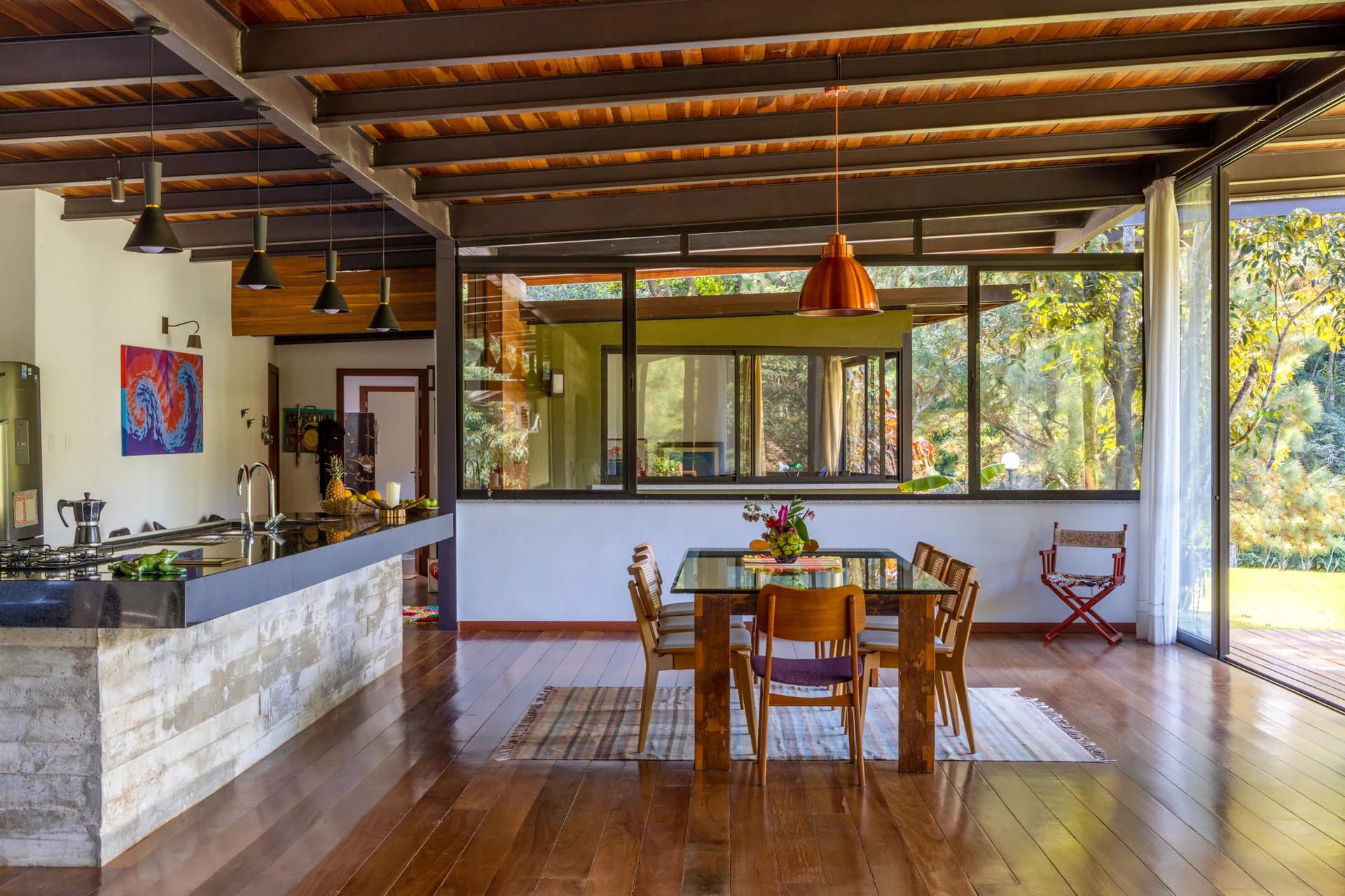 A modern dining area features a glass-top table, wooden chairs, and a kitchen island under an exposed steel beam and wood plank ceiling.