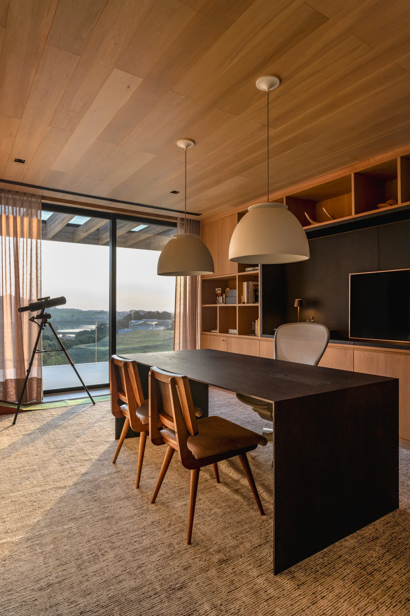 A modern home office with a dark desk, dome pendant lights, built-in wooden shelving, and a glass wall showing an outdoor landscape.