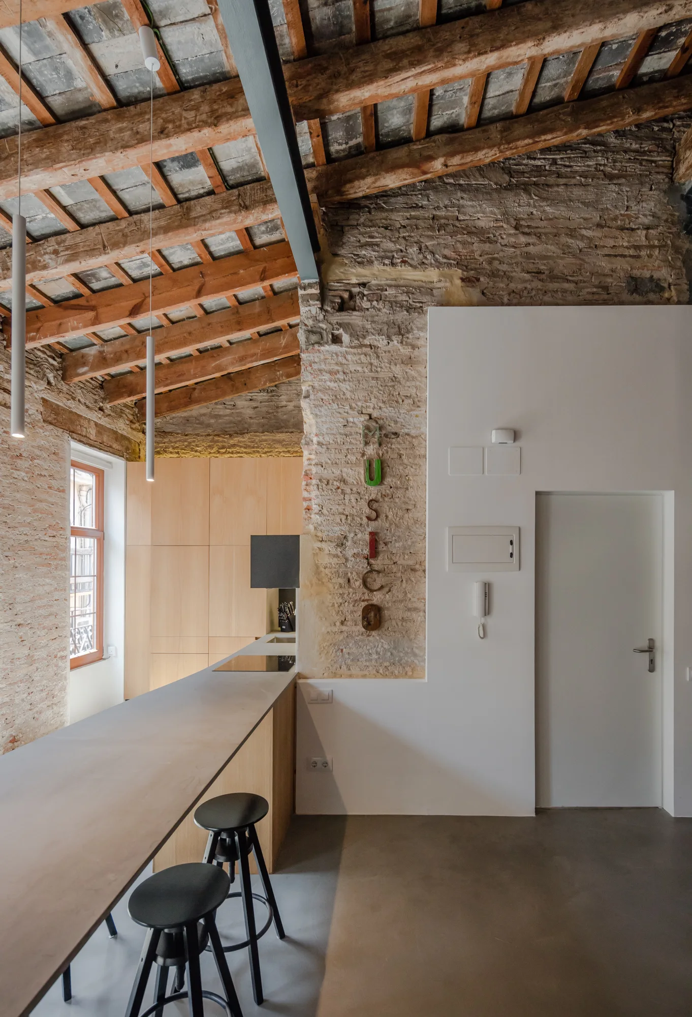A modern kitchen features a long concrete-look breakfast bar with black stools, set against exposed brick walls and a rustic timber ceiling.