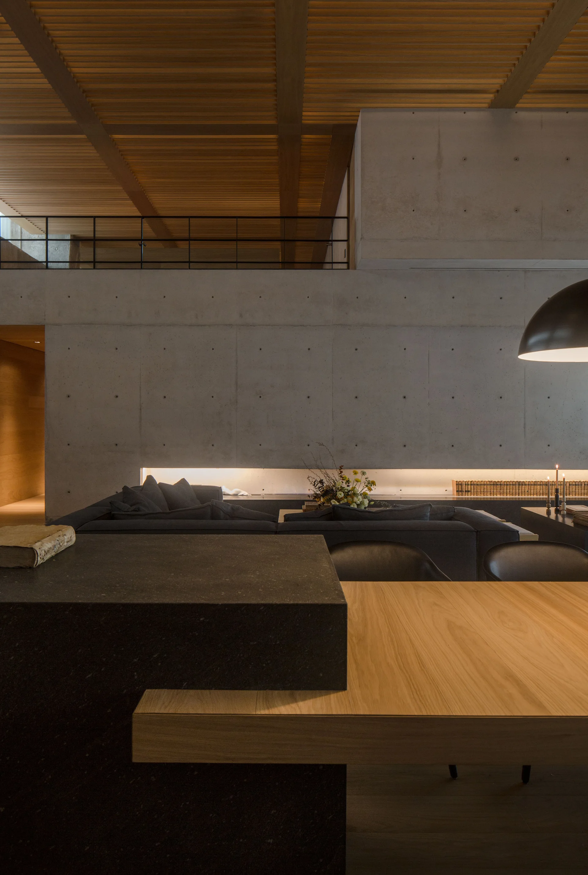 A modern living area with a large exposed concrete wall, a slatted wood ceiling, a dark sectional sofa, and a wooden dining table.