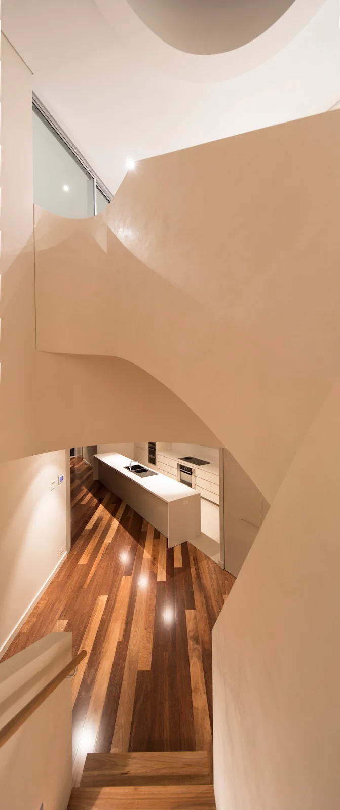 High-angle view from a curved beige staircase looking down at a minimalist white kitchen island and glossy hardwood flooring.