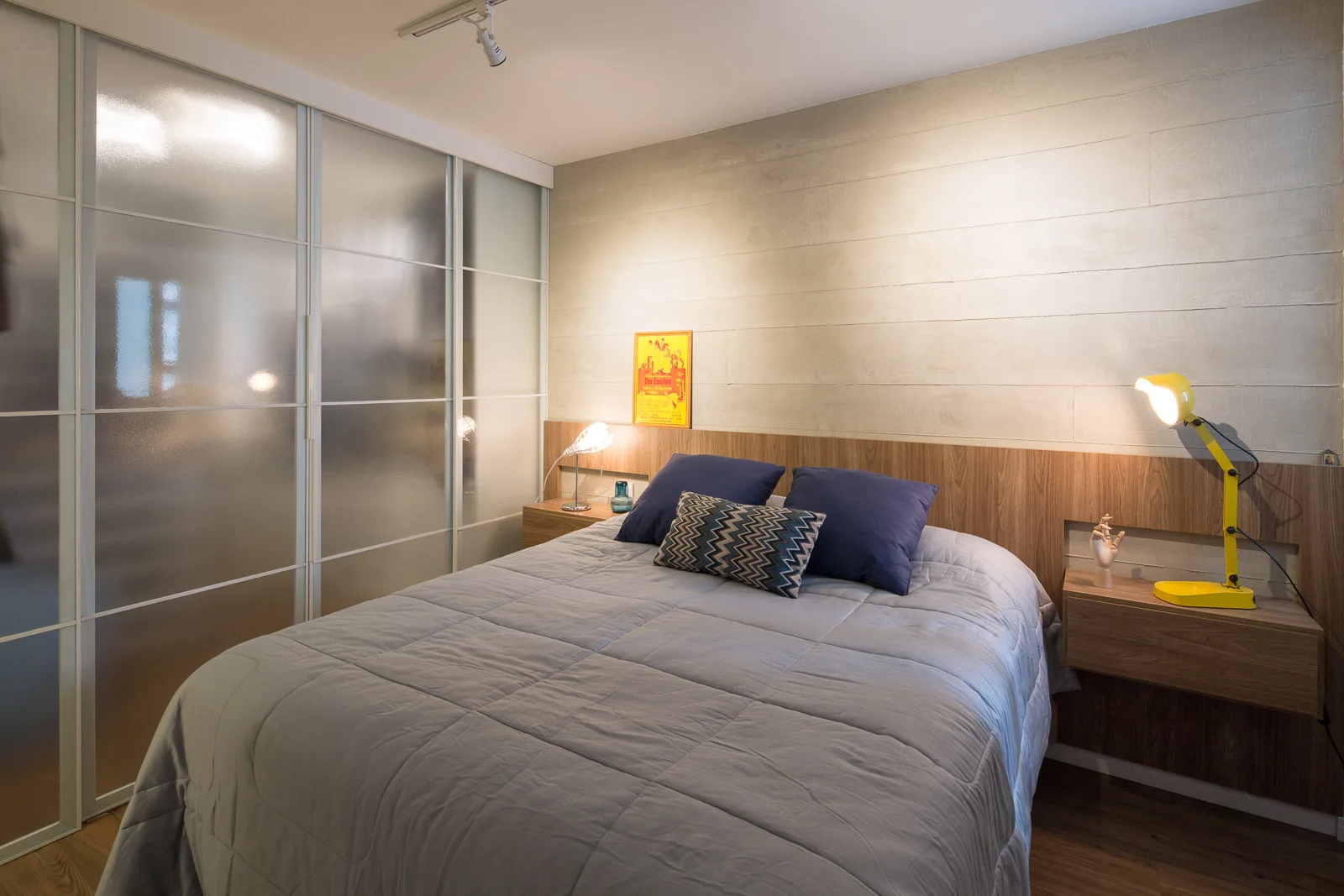 A modern bedroom features a grey bed between floating wooden nightstands set against a concrete wall and frosted glass sliding doors.