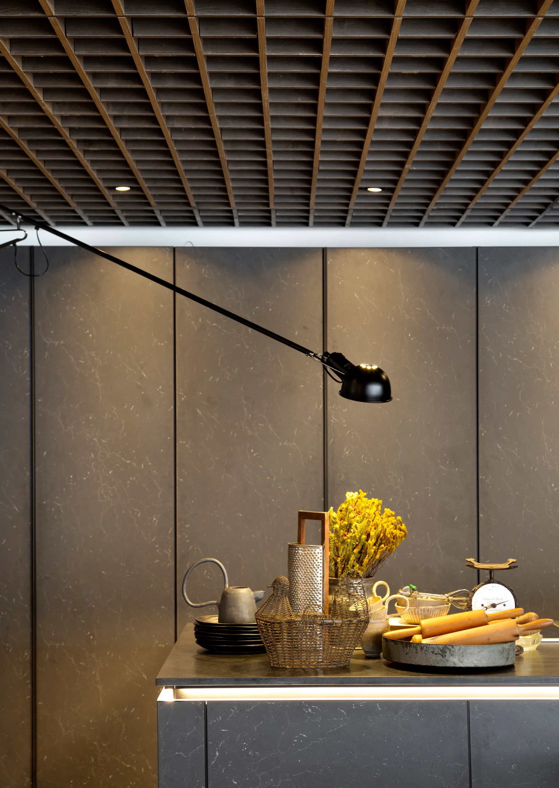 A dark charcoal kitchen island with cooking tools sits below a wooden grid ceiling and a black adjustable swing-arm lamp mounted on a paneled wall.