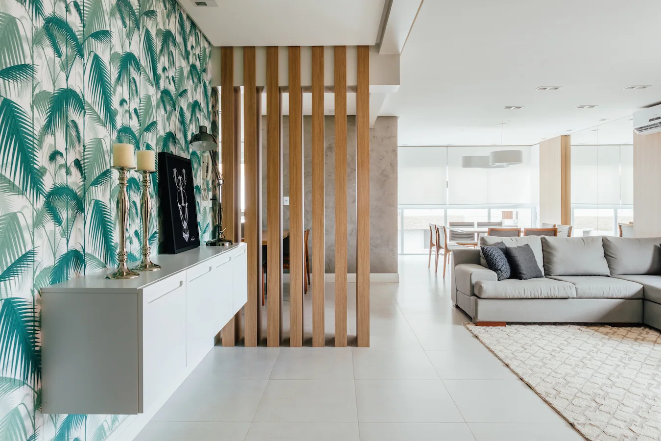 A modern living space features a green palm leaf wallpaper accent wall, a white floating cabinet, and a vertical wood slat room divider.