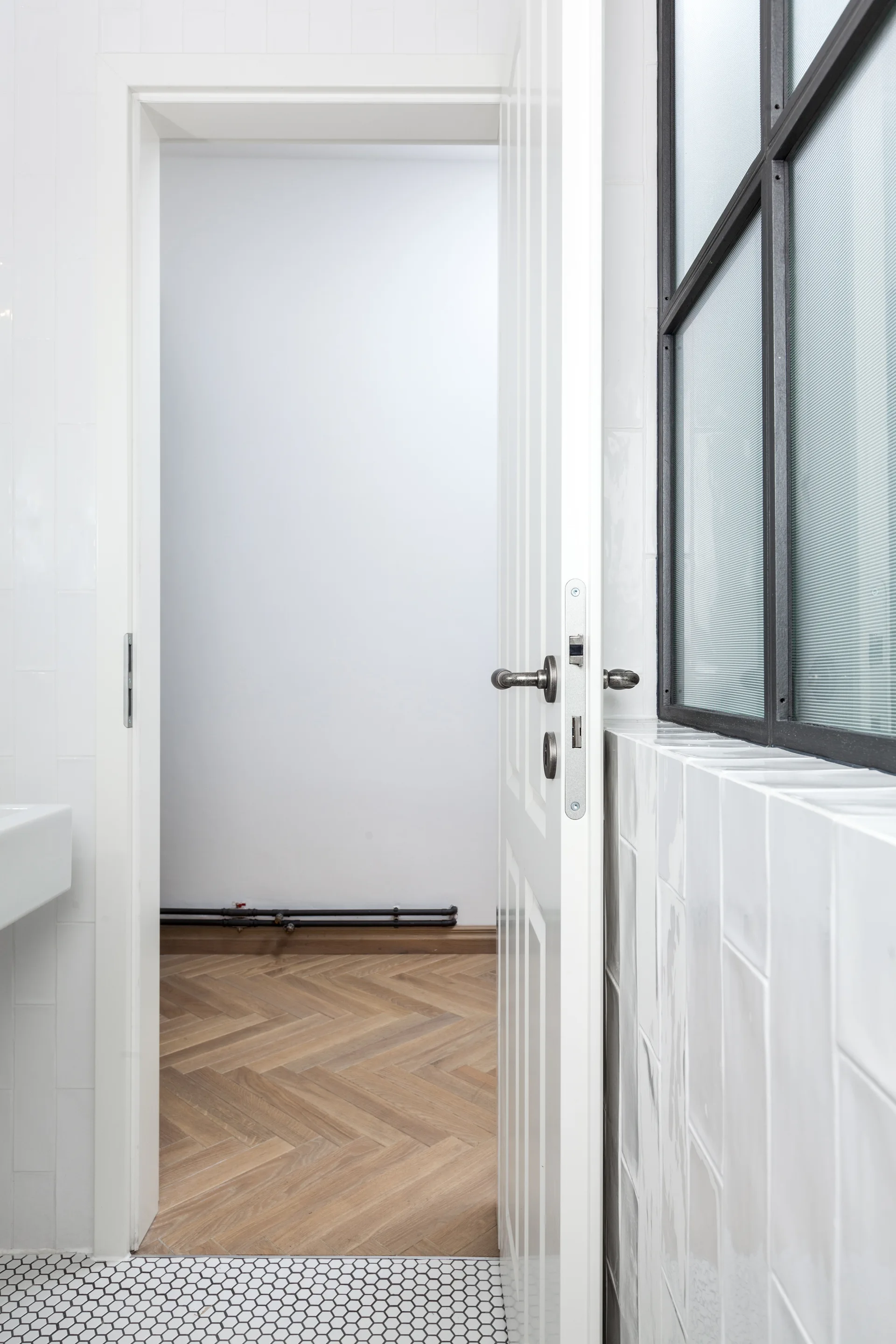 An open white door connects a bathroom with hexagon floor tiles and vertical wall tiles to a hallway with herringbone wood floors.
