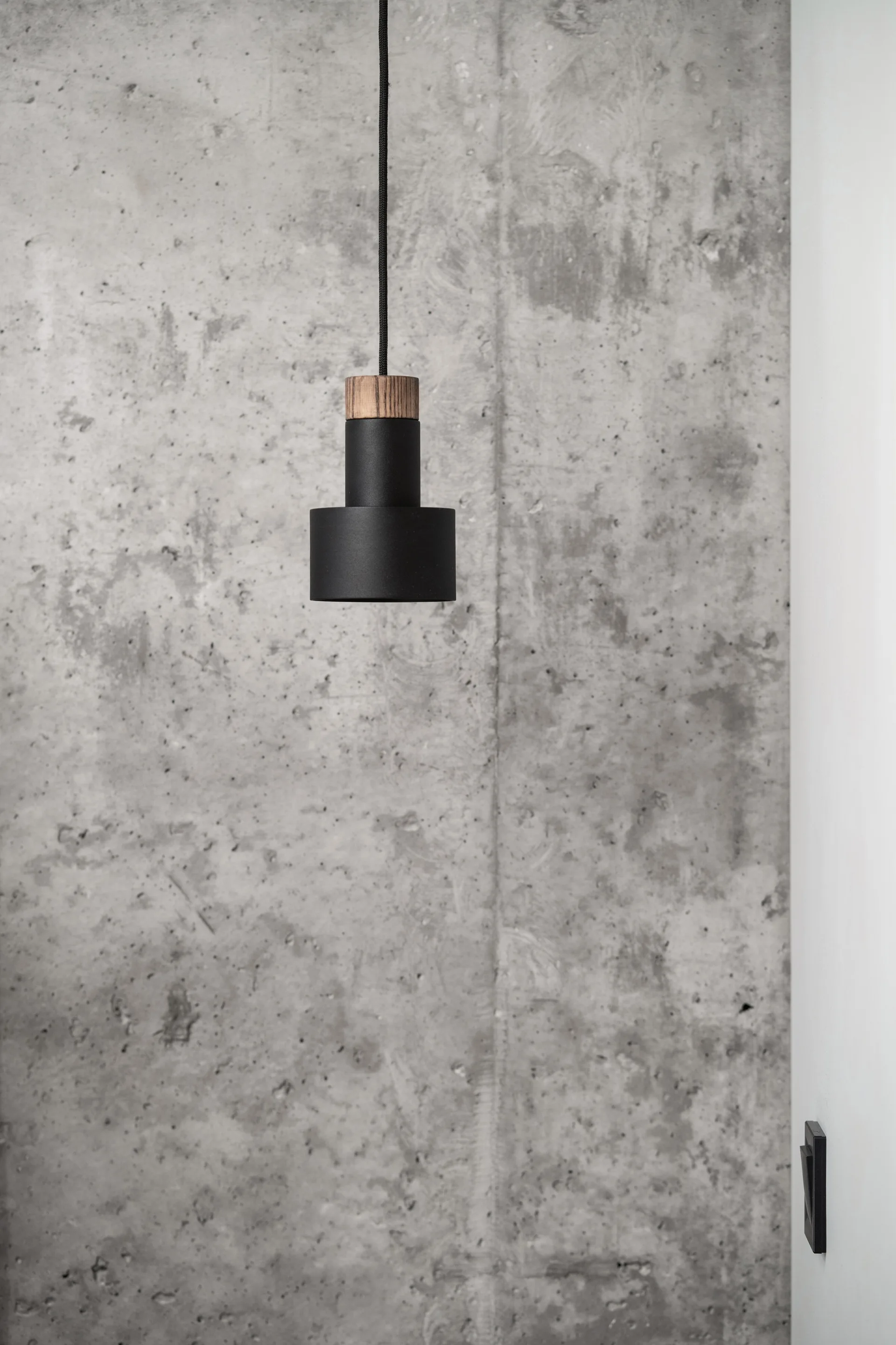 A matte black cylindrical pendant light with a wood collar hangs in front of a textured grey concrete wall.
