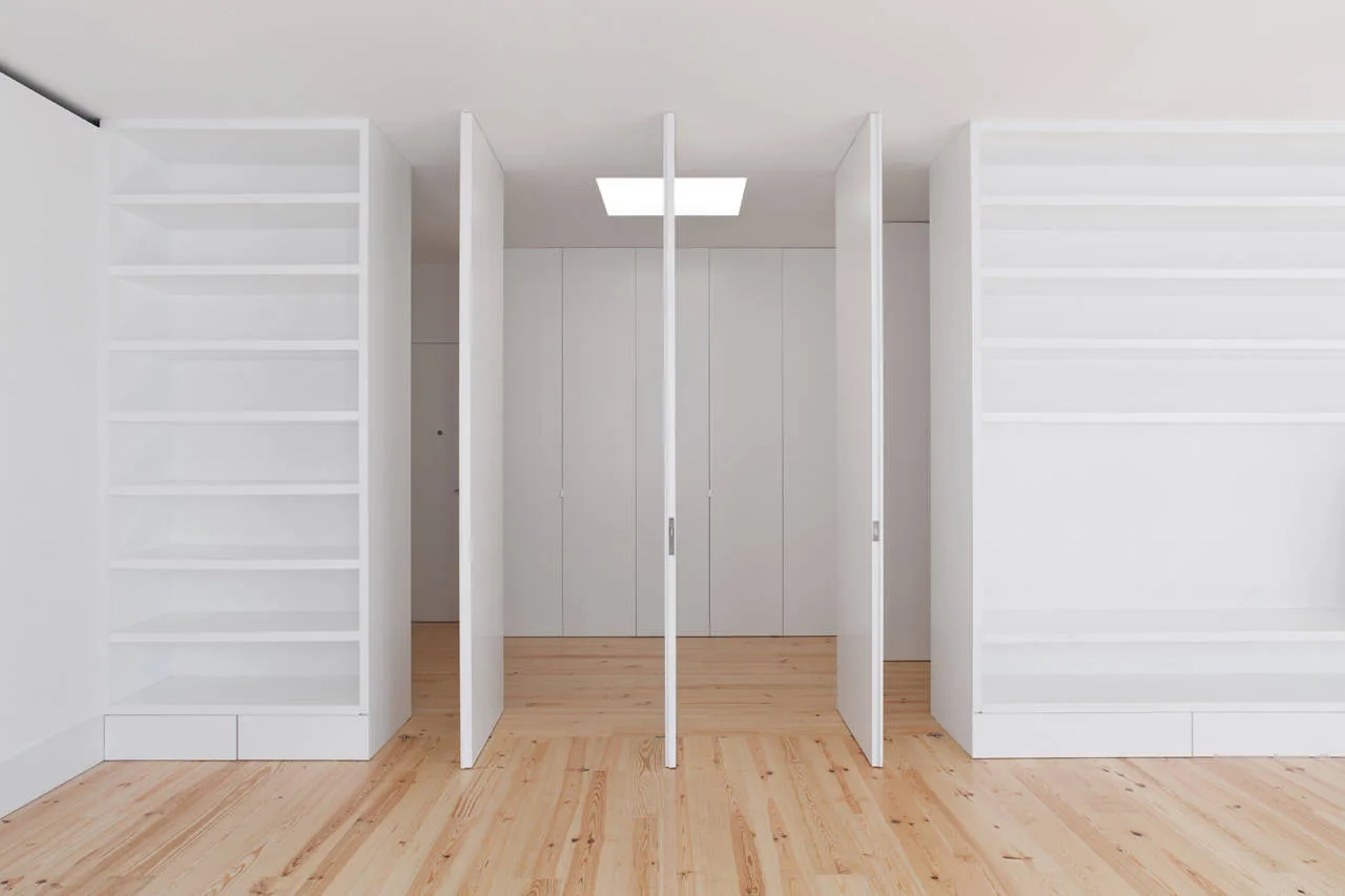 Minimalist white built-in bookshelves frame tall pivoting white doors in an interior hallway featuring natural light-colored wood plank flooring.