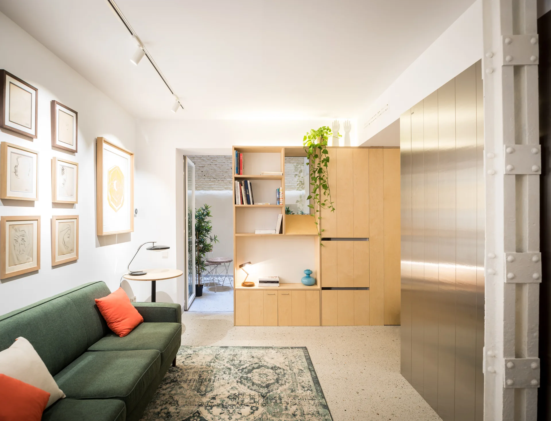 A contemporary living room featuring a green sofa, patterned rug, light wood built-in cabinetry, a brushed metal wall panel, and terrazzo flooring.