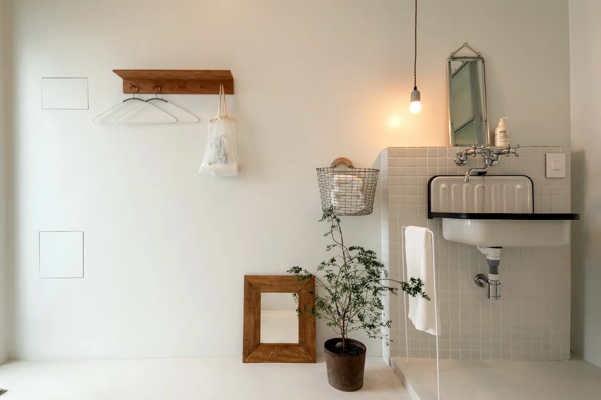 A minimalist washroom features a wall-mounted vintage sink against white grid tiles, a hanging bulb light, and wooden decor accents.