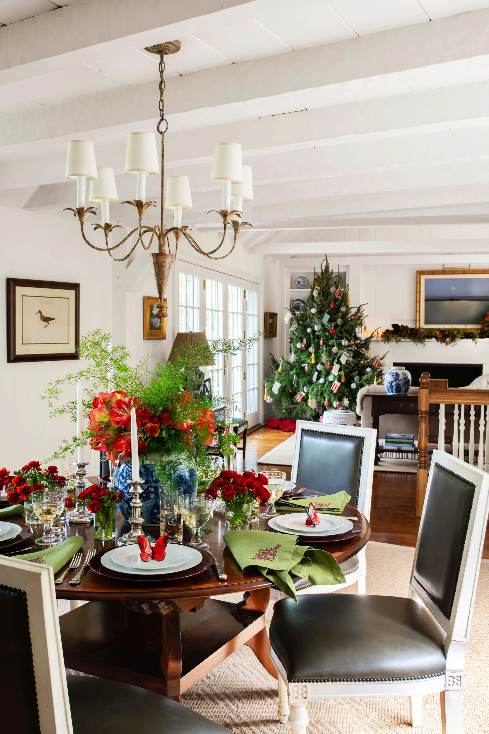 A festive dining room featuring a round wooden table set with red flowers and green napkins beneath a white beamed ceiling and metal chandelier.