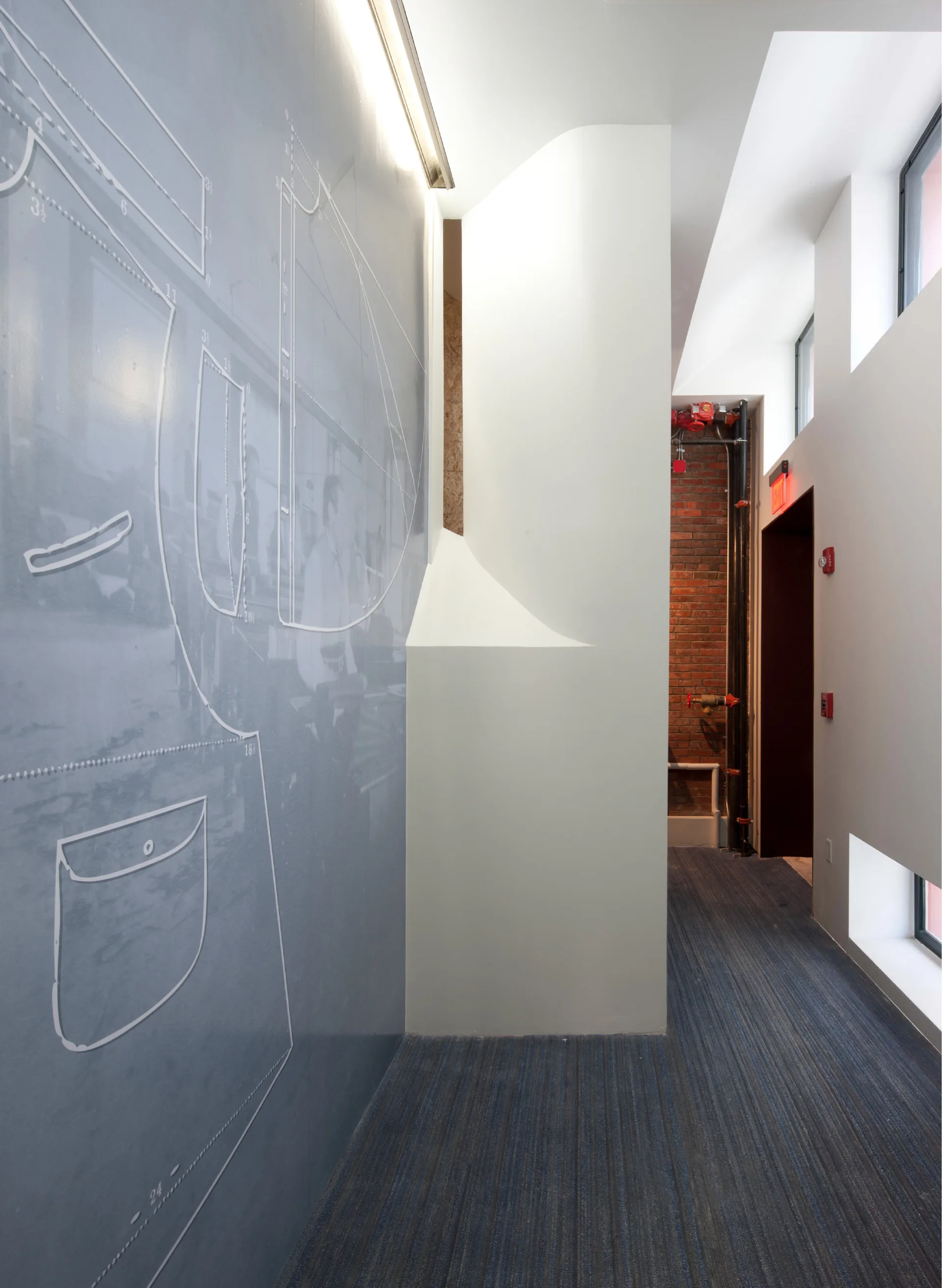 A modern hallway featuring a blue wall mural with white schematic drawings, striped carpet, sculptural white drywall, and an exposed brick accent wall.