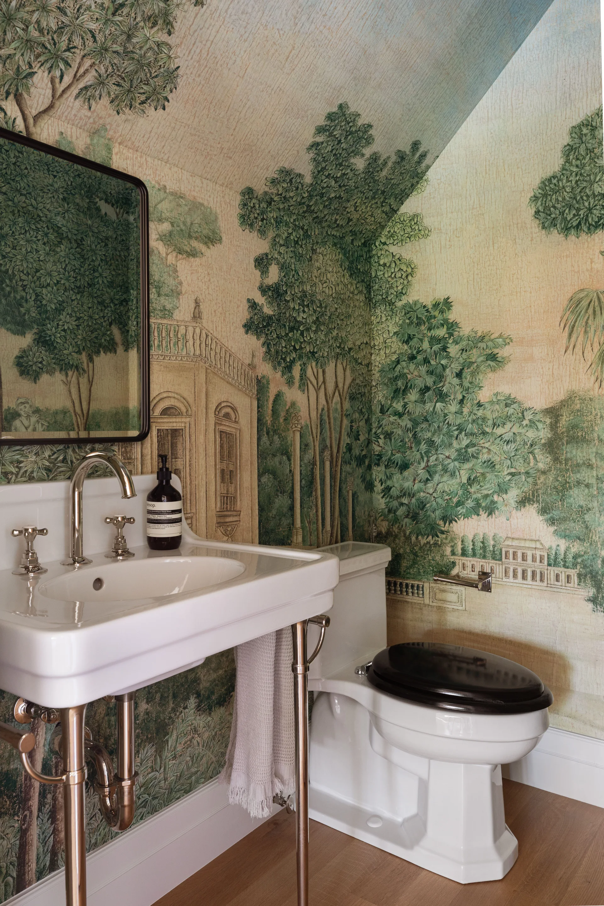 A powder room features green and beige scenic mural wallpaper behind a white console sink with metal legs and a white toilet with a dark wood seat.