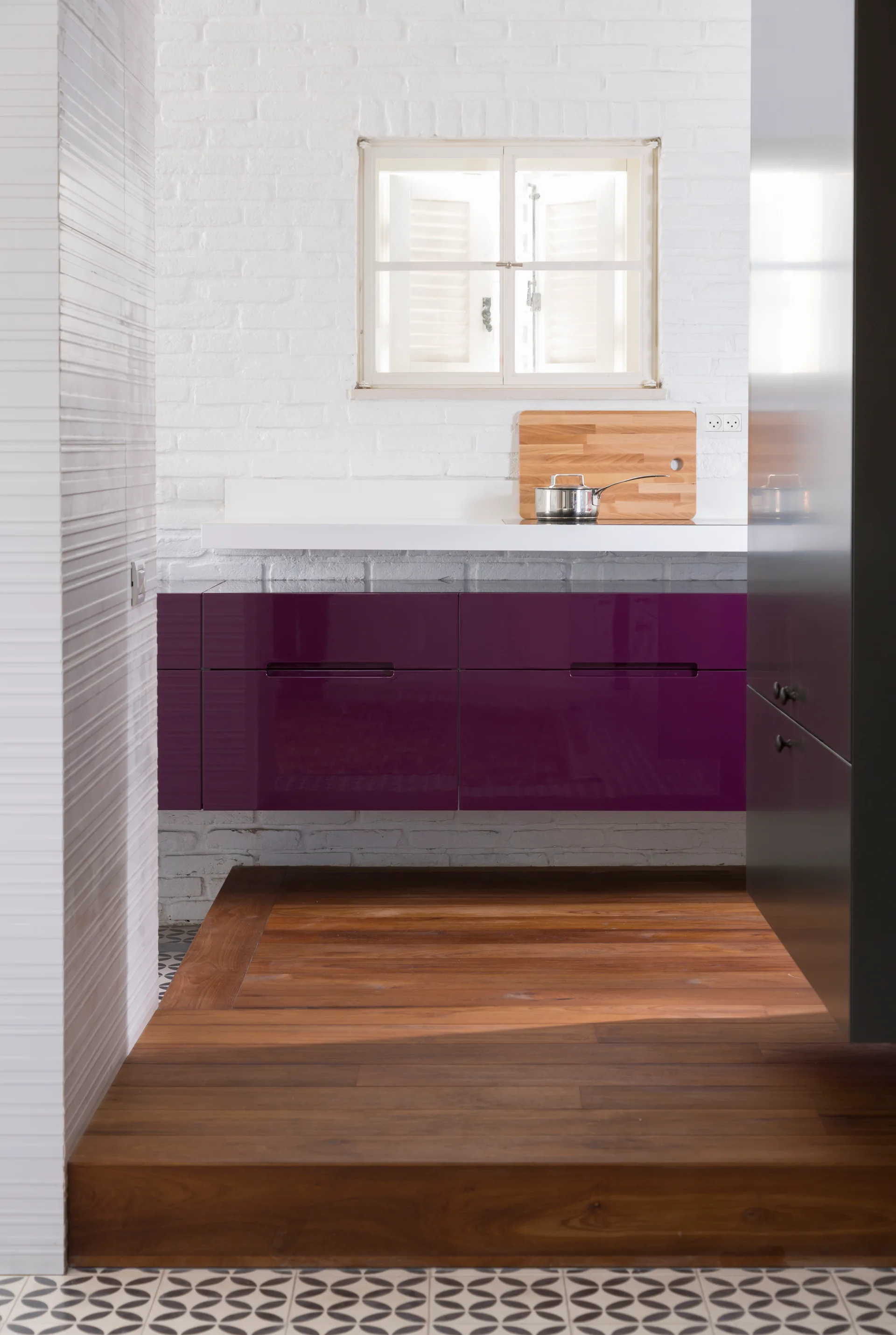 A modern kitchen with glossy purple floating cabinets, a white painted brick wall with a small window, and a raised wood floor transition to patterned tile.