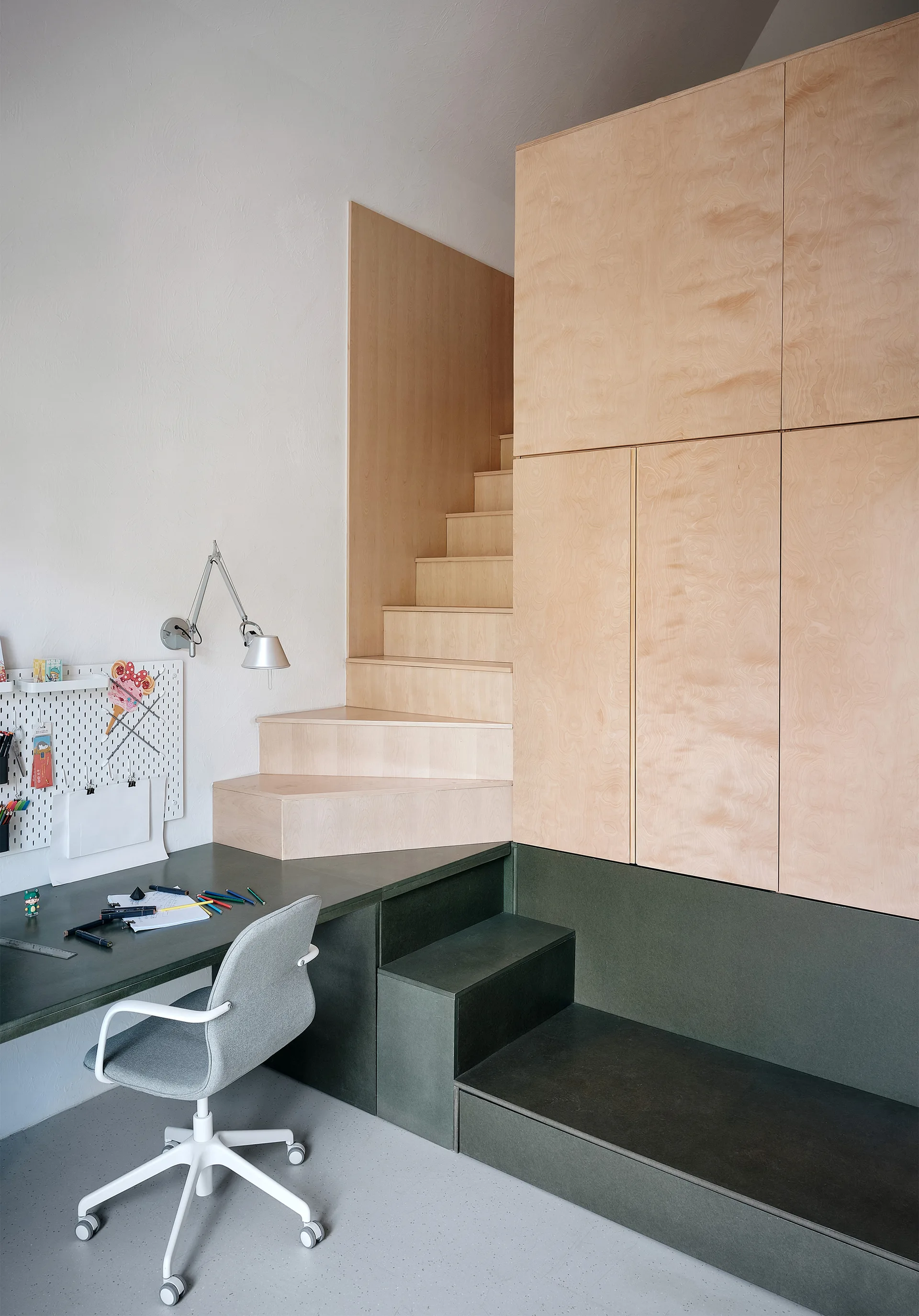 A compact home office workspace featuring a dark green built-in desk, a grey task chair, and a light wood plywood cabinet unit with integrated stairs.