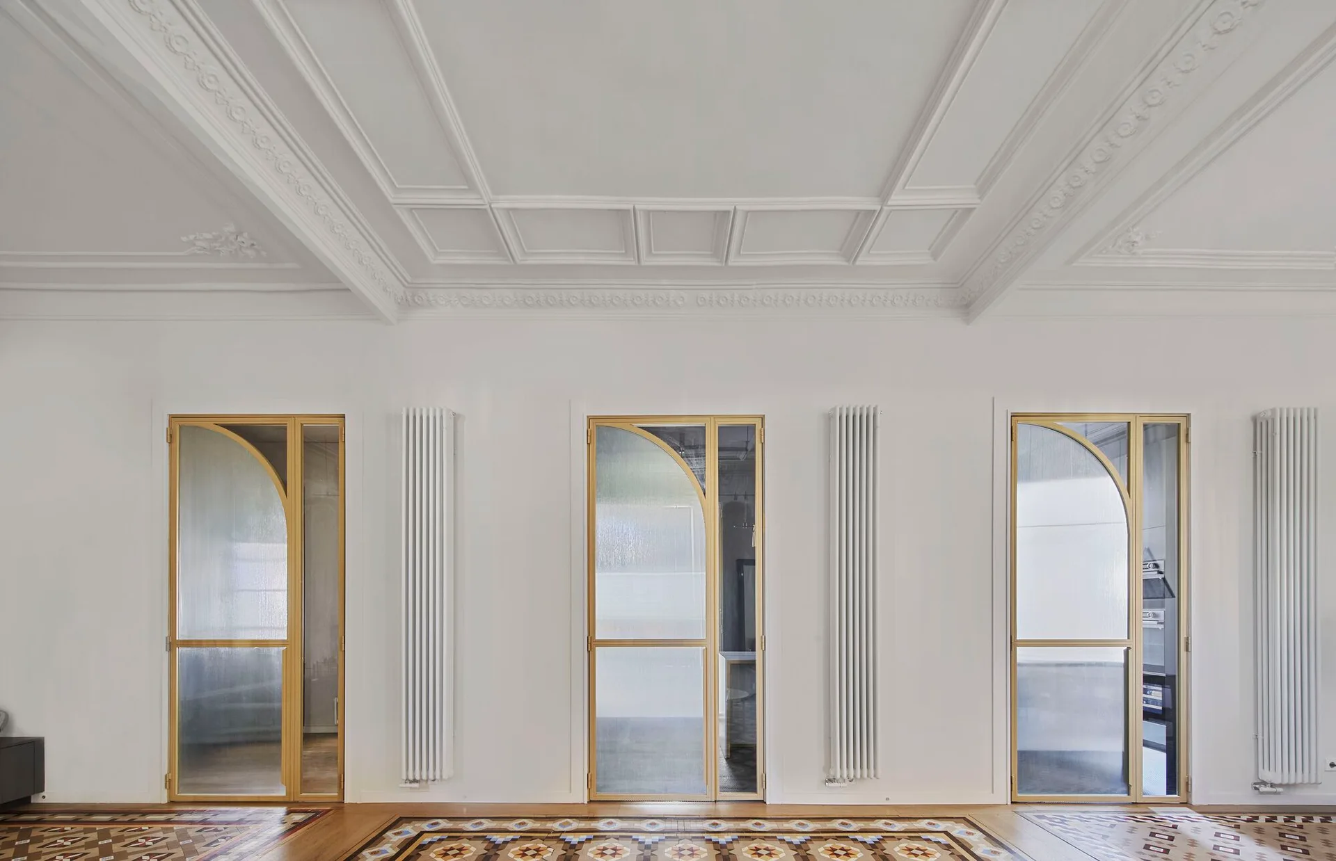 Three gold-framed doors with glass arch inserts stand between vertical white radiators below an ornate coffered ceiling and a patterned floor.