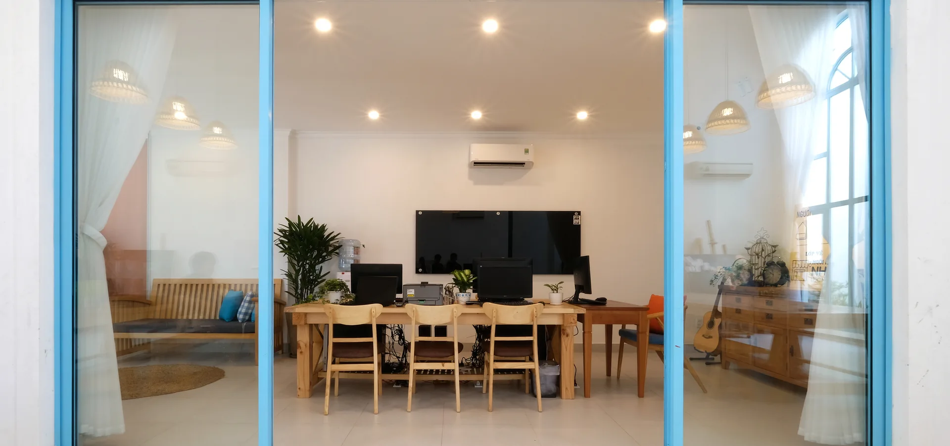 A home office setup with a long wooden desk and chairs viewed through a sliding glass door with a bright blue frame.
