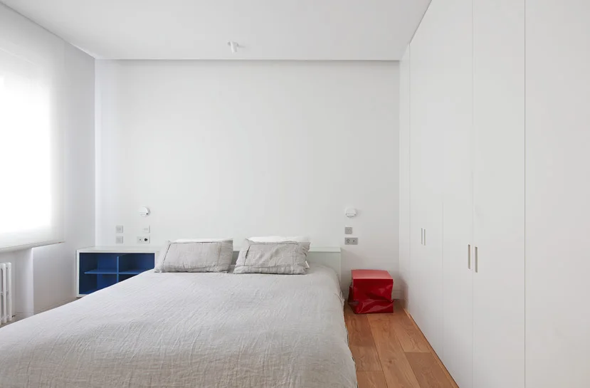 A minimalist white bedroom features a bed with grey linen bedding, a blue open nightstand, a red stool, and a built-in wardrobe.