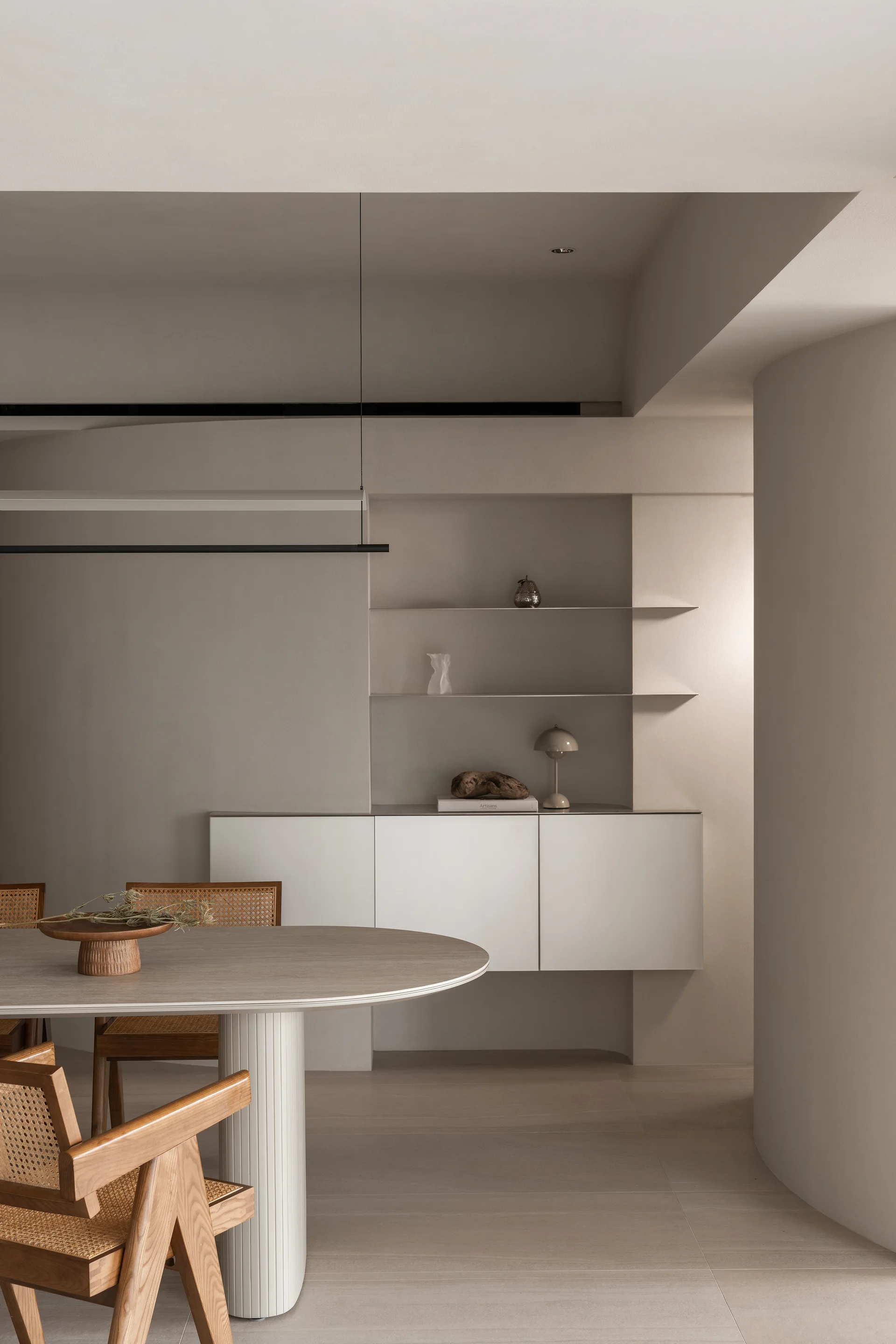 A minimalist dining room features a light wood table with a fluted pedestal, woven cane chairs, and recessed niche shelving against a beige wall.