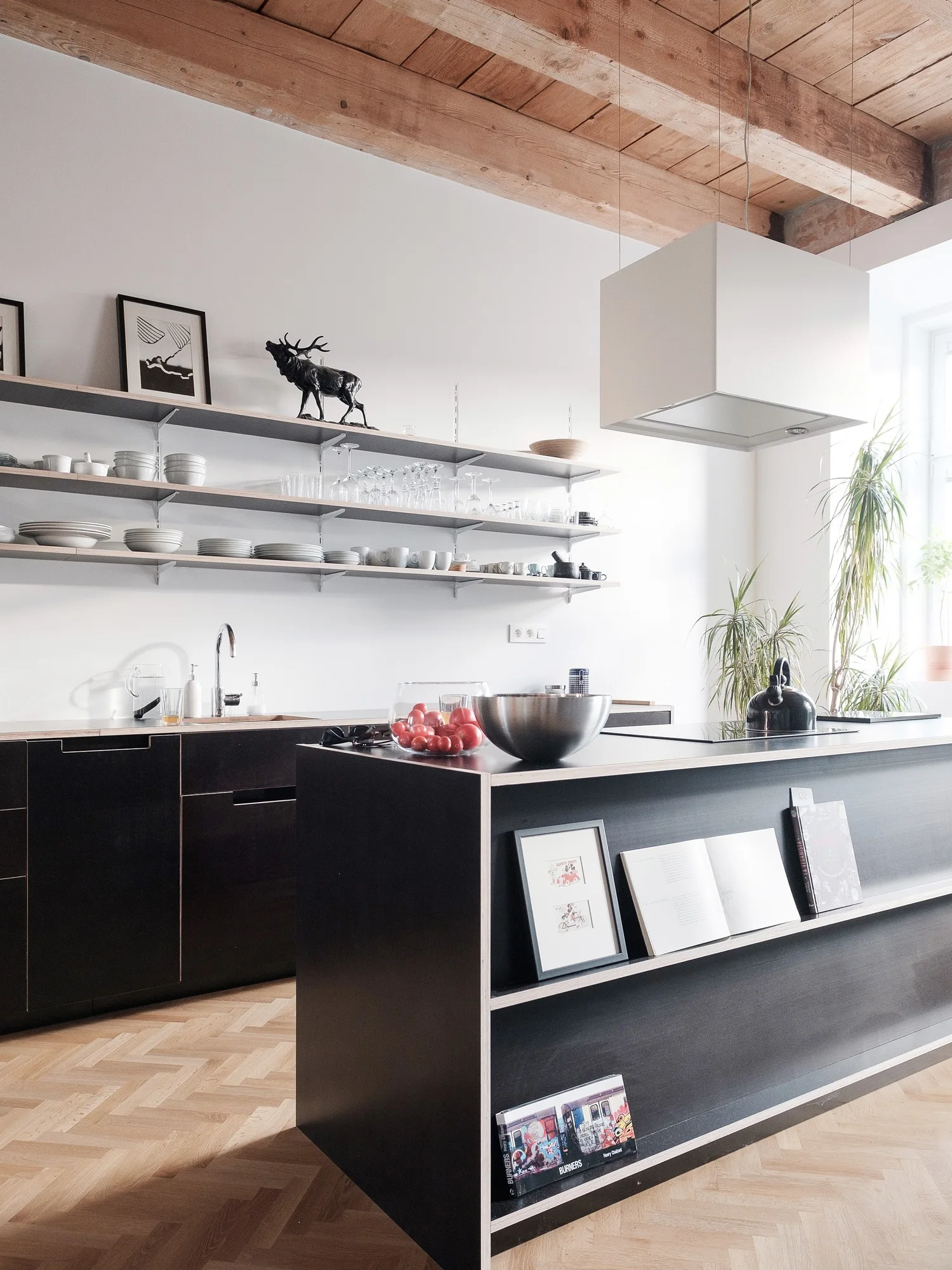 A modern kitchen featuring black lower cabinets with exposed plywood edges, open wall shelving, herringbone wood floors, and exposed ceiling beams.