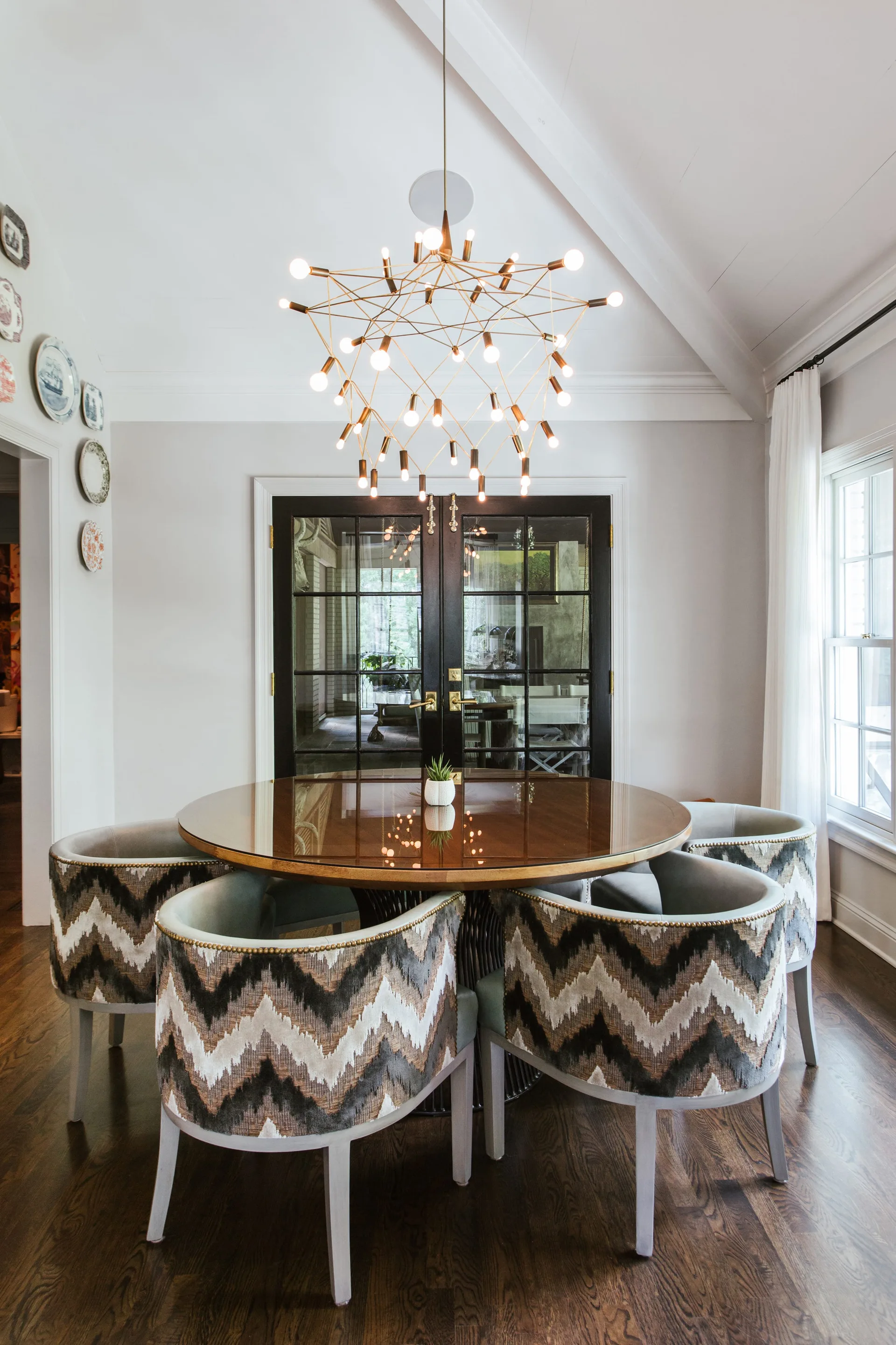 A round wooden dining table surrounded by grey and brown chevron patterned chairs under a large gold sputnik chandelier.