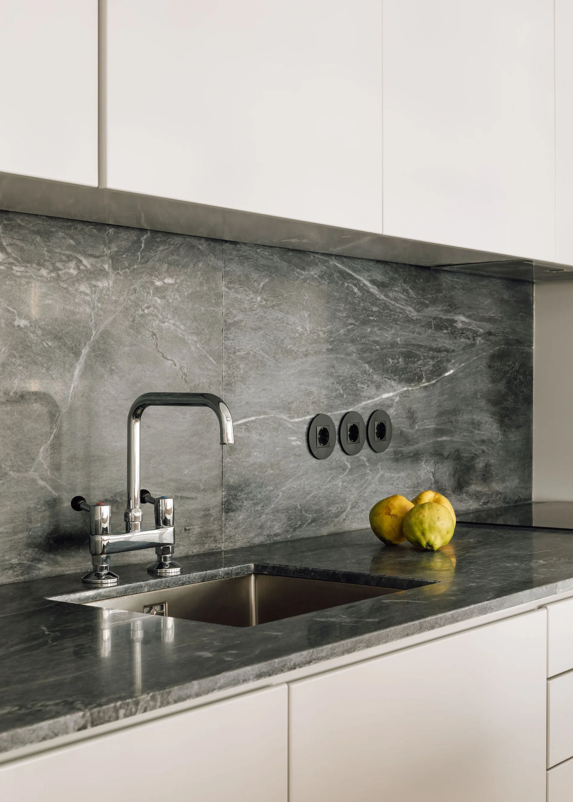 A close-up of a kitchen sink with a chrome faucet set in a grey marbled stone countertop and matching backsplash beneath white flat-panel cabinets.