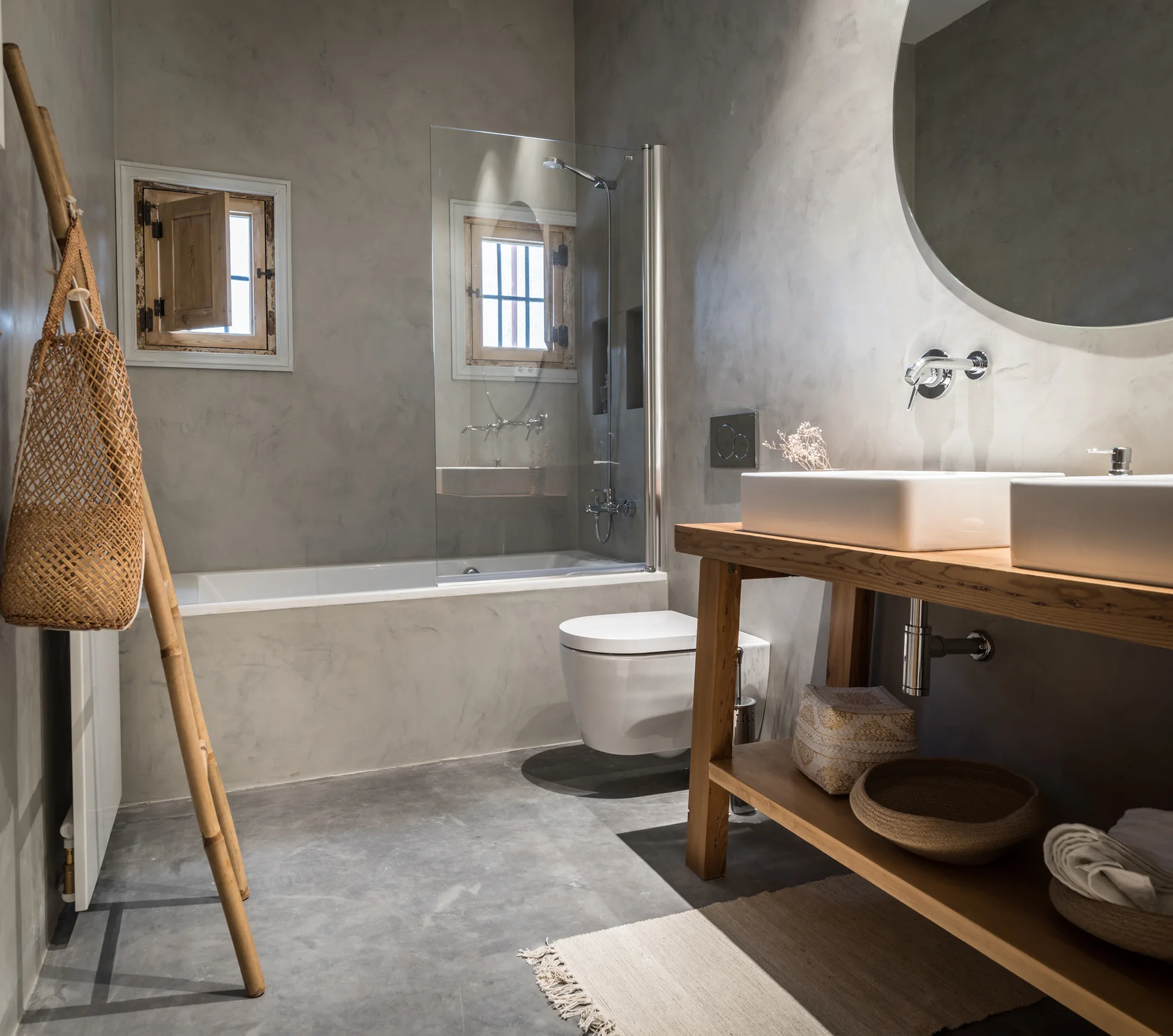A minimalist bathroom features grey plaster walls, a natural wood open vanity with two white sinks, a bathtub, and a wall-mounted toilet.