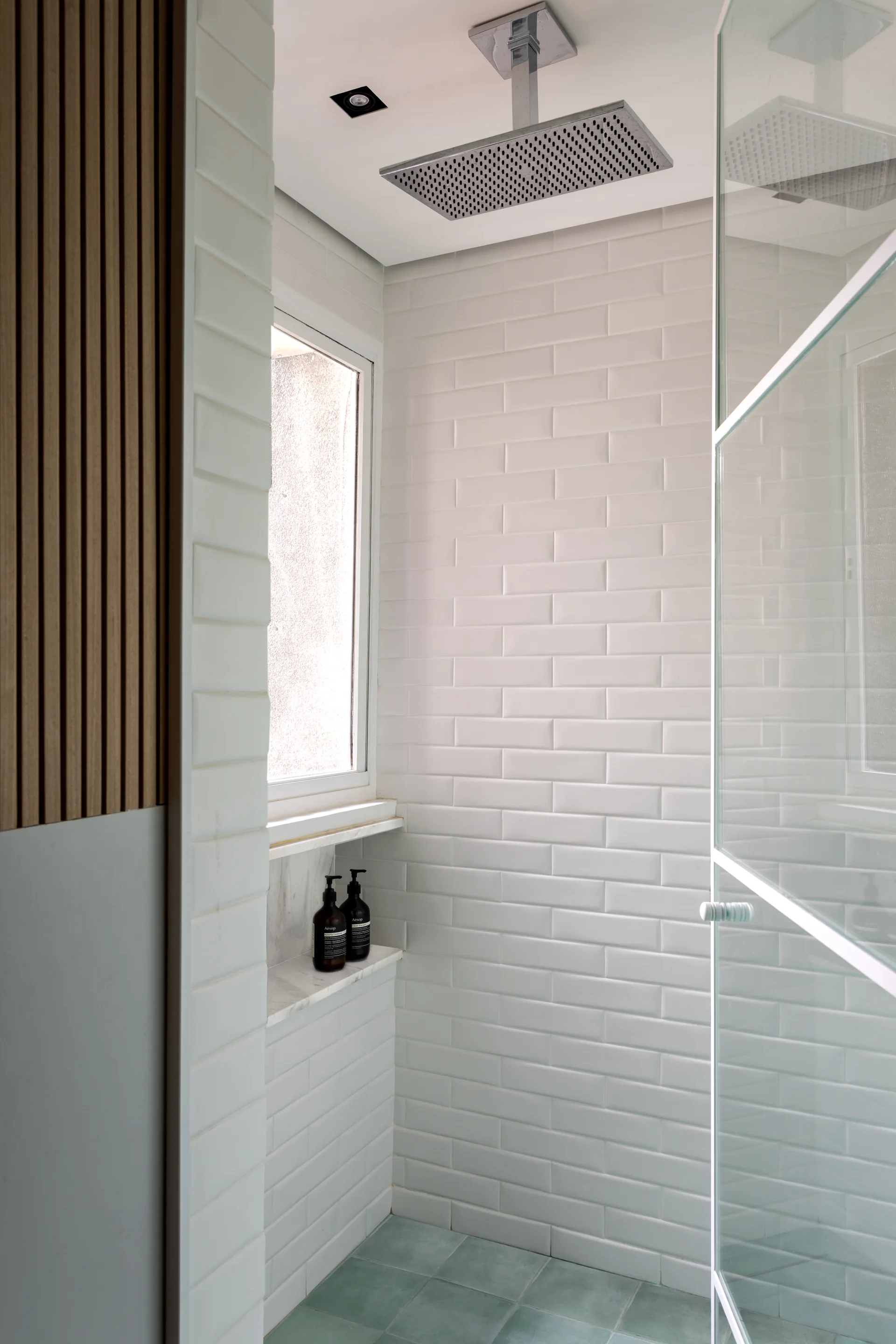A modern bathroom shower features white subway tiled walls, a ceiling-mounted rain shower head, pale green floor tiles, and a vertical wood slat accent panel.