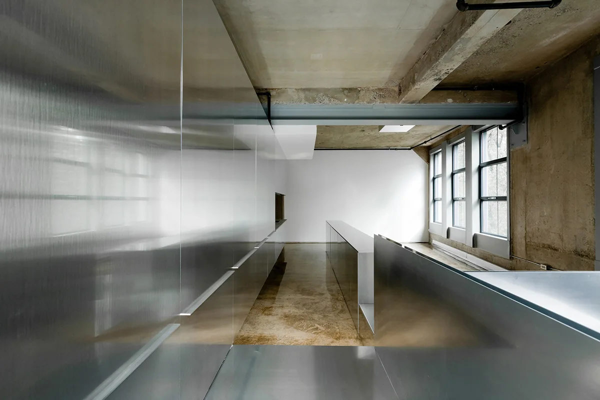 A modern industrial corridor features full-height brushed stainless steel cabinetry, a raw concrete ceiling, and large rectangular windows.
