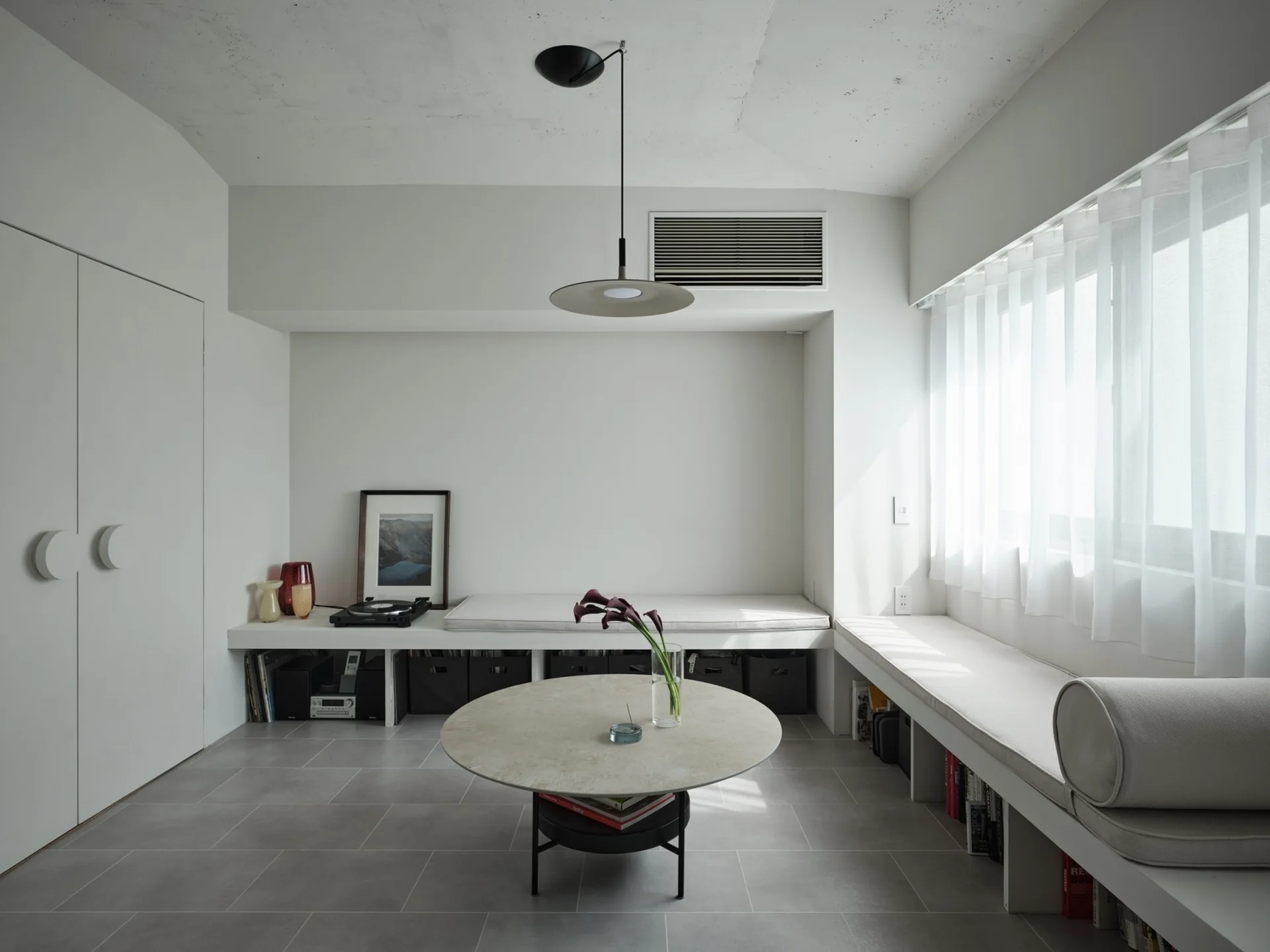A minimalist living area features low built-in white benches with grey cushions, a round stone-topped coffee table, and large-format grey tile flooring.