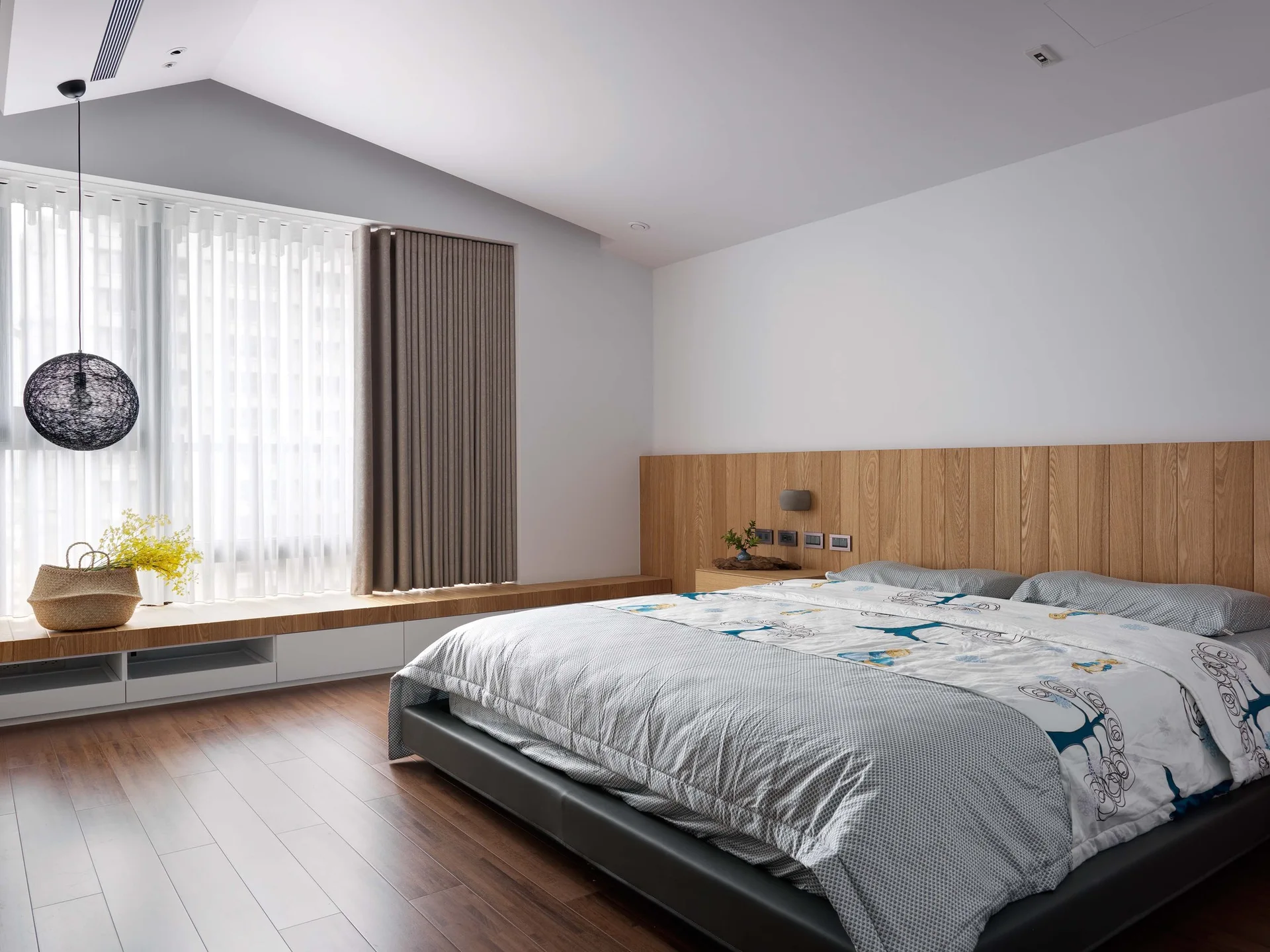 A modern bedroom featuring a low-profile bed with grey patterned bedding, a wood-paneled accent wall, and a long built-in window bench.