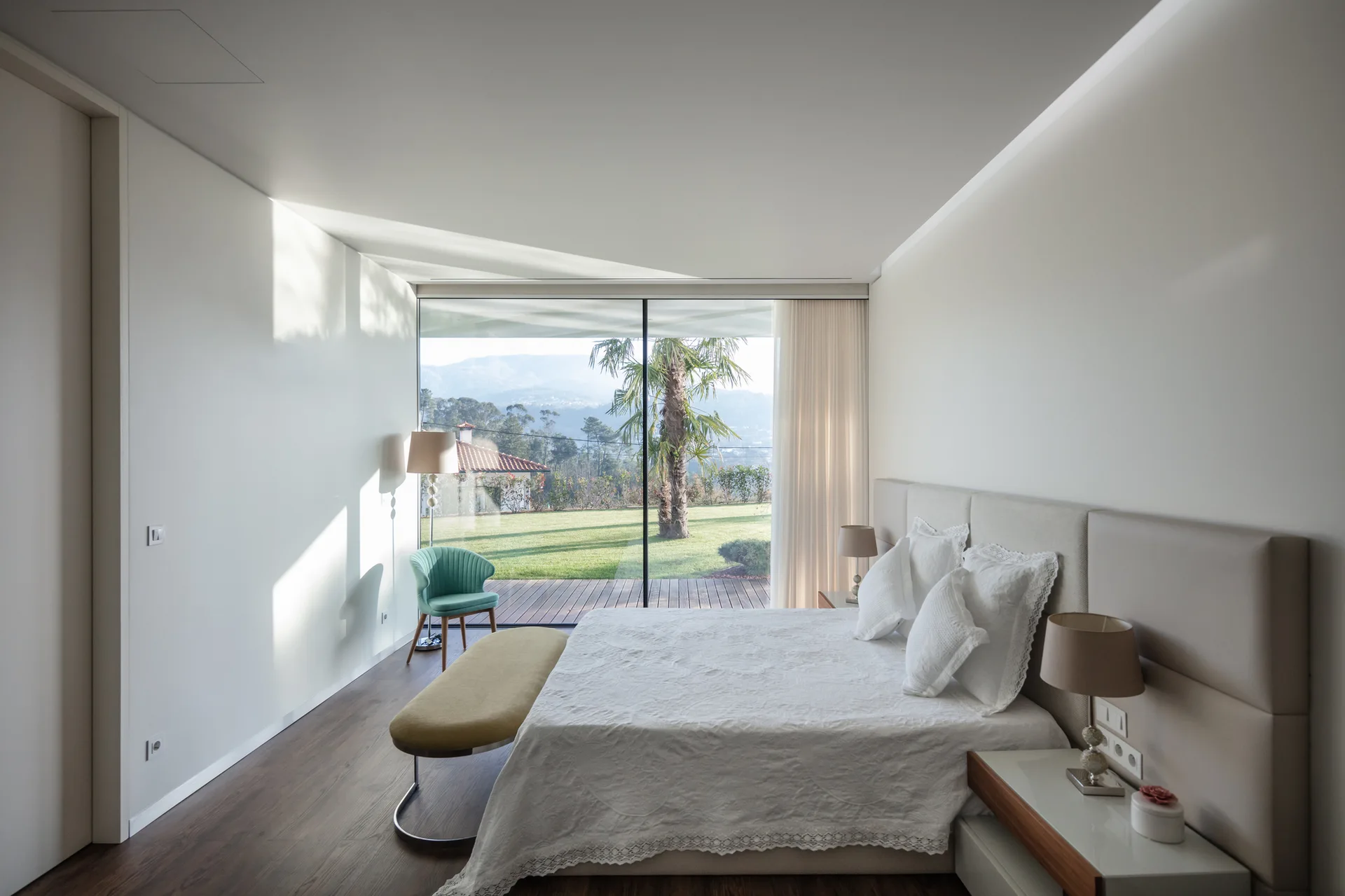 A modern bedroom features a large bed with a beige headboard, built-in nightstands, a teal armchair, and sliding glass doors opening to a grassy lawn.