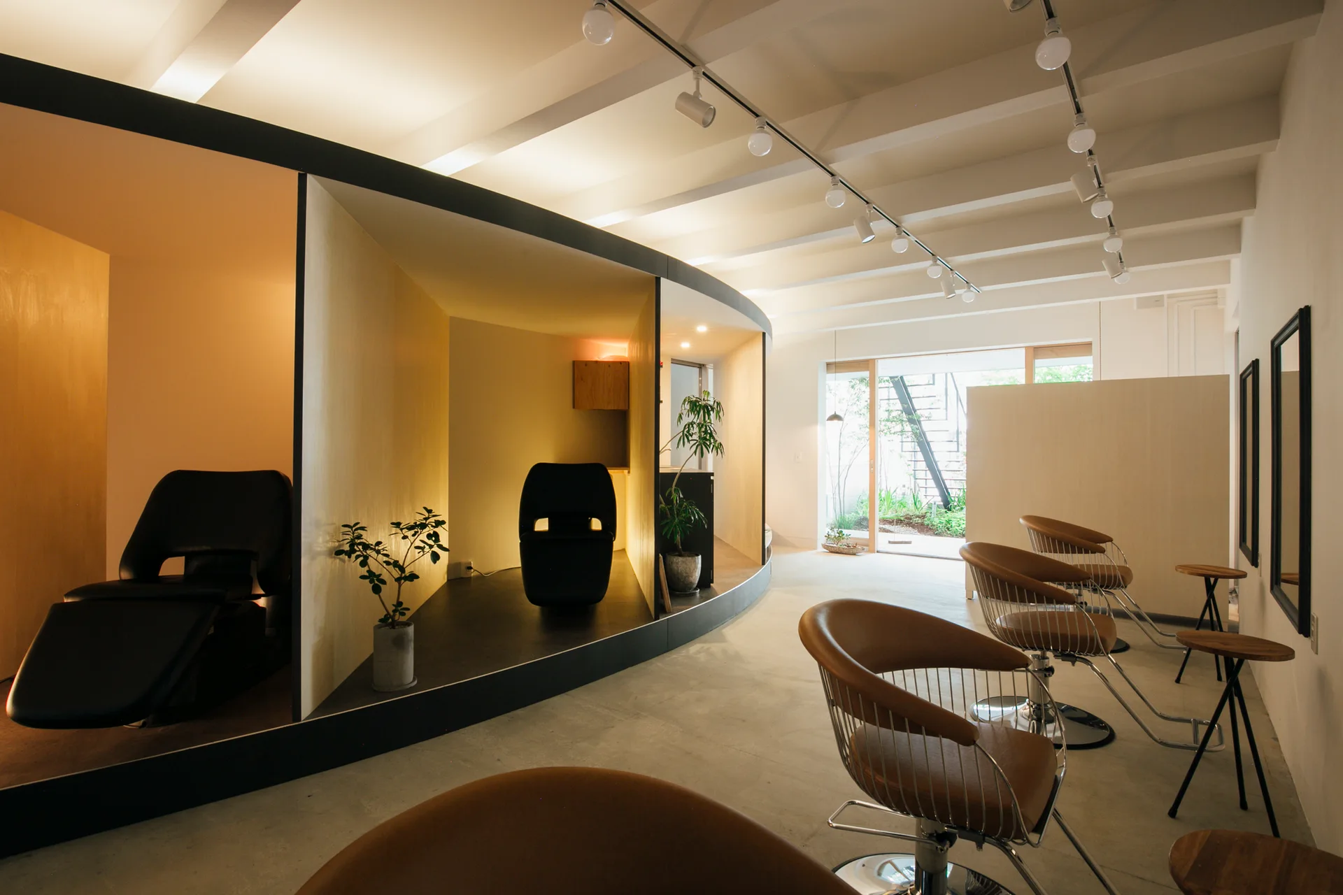A modern salon interior featuring a curved light wood partition wall housing treatment chairs, alongside minimalist brown styling chairs on a concrete floor.